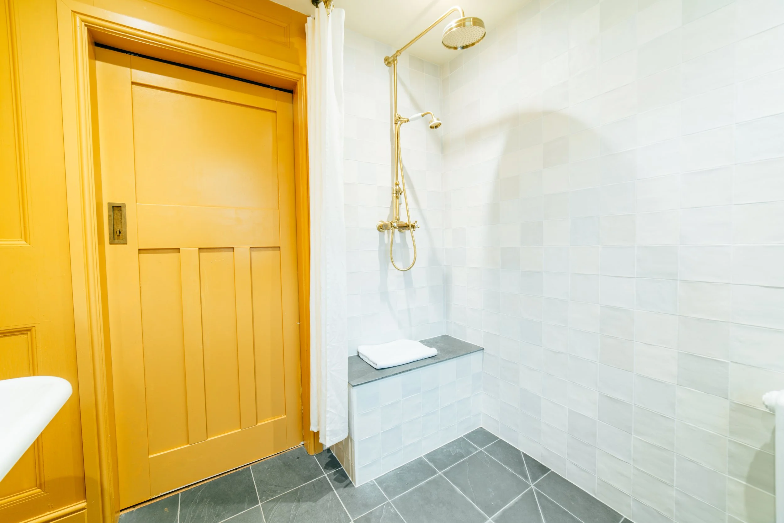 Empty walk-in shower with white tiled walls, gray tiled floor, a brass showerhead, and a folded towel on a small seat.