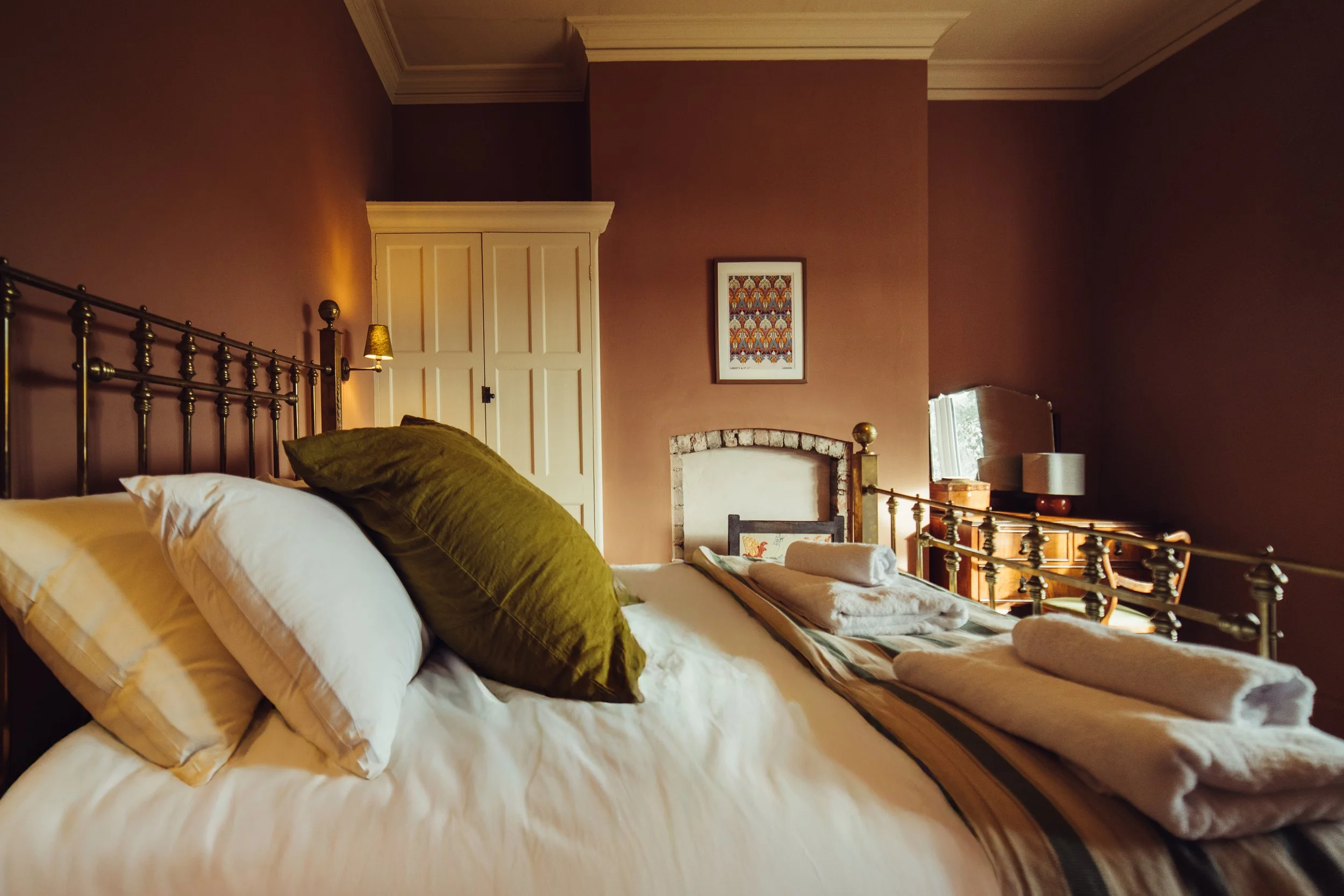 A cozy bedroom with a large bed that has white and green pillows, rolled towels, in a warm-colored room with a cabinet, a small mirror, and a painting on the wall.