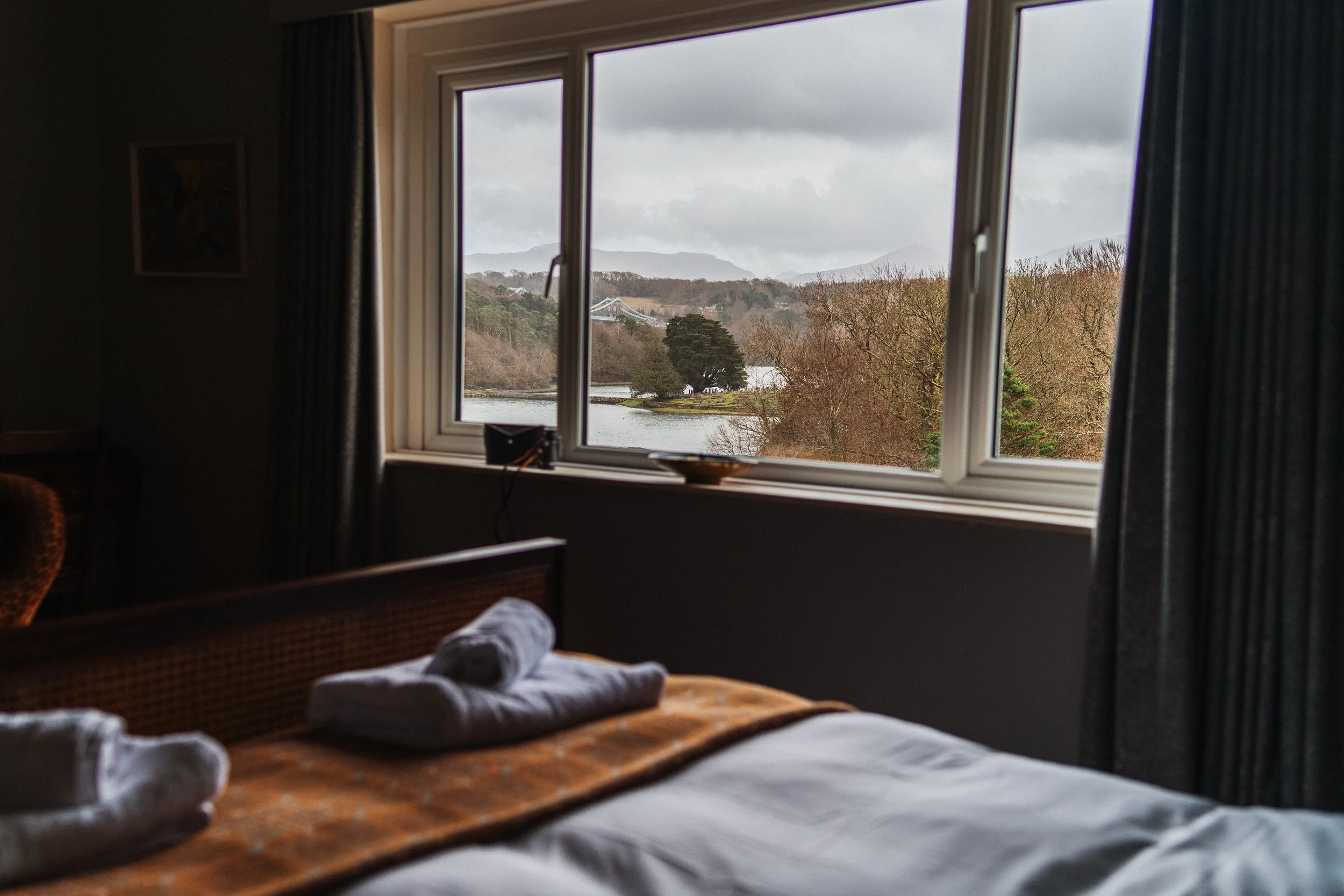 A view from a bedroom window showing a river, trees, and mountains in the distance on a cloudy day.