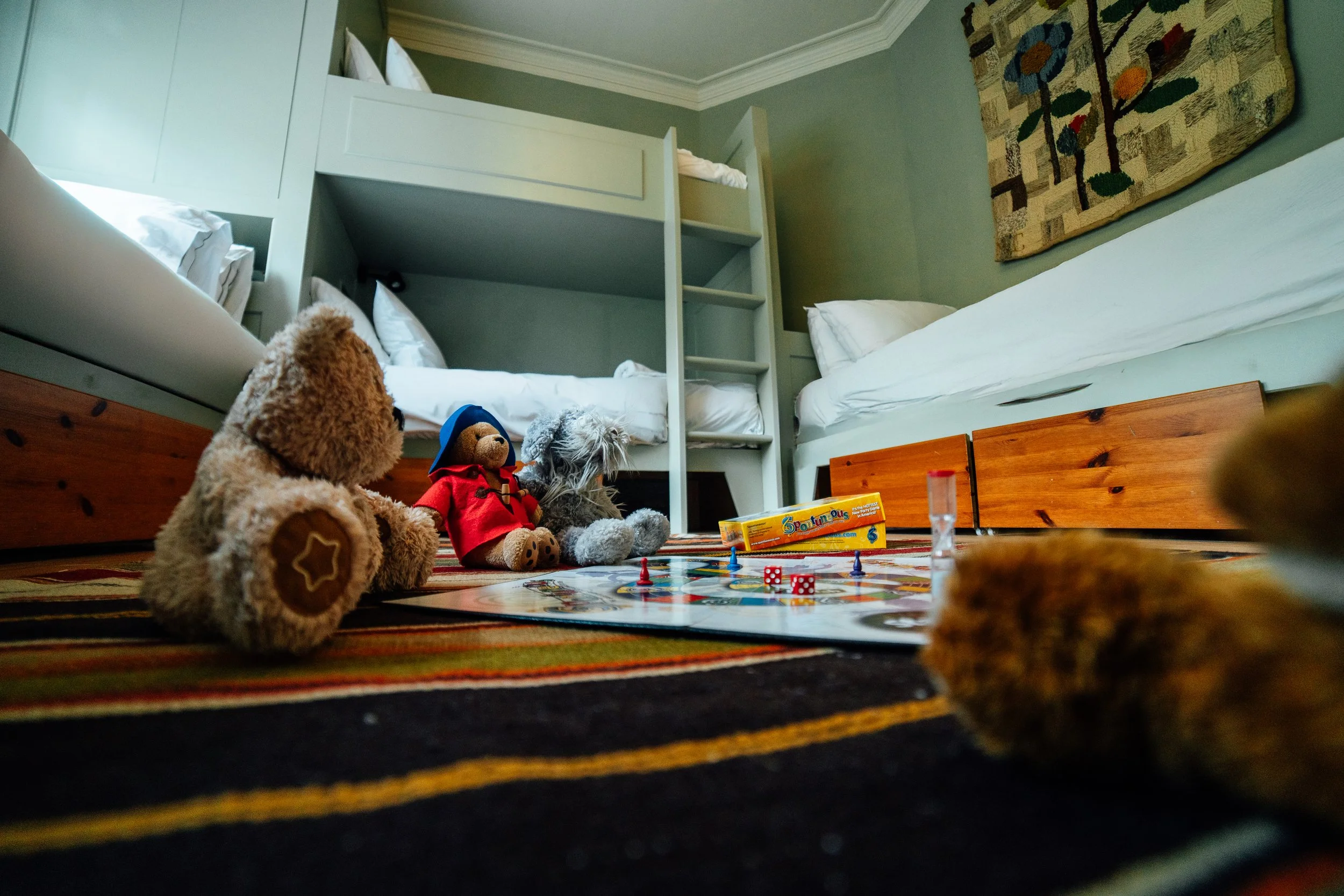 Children's toys, including stuffed animals and board game pieces, are scattered on a striped rug in a bedroom, near a bunk bed and white bedding.