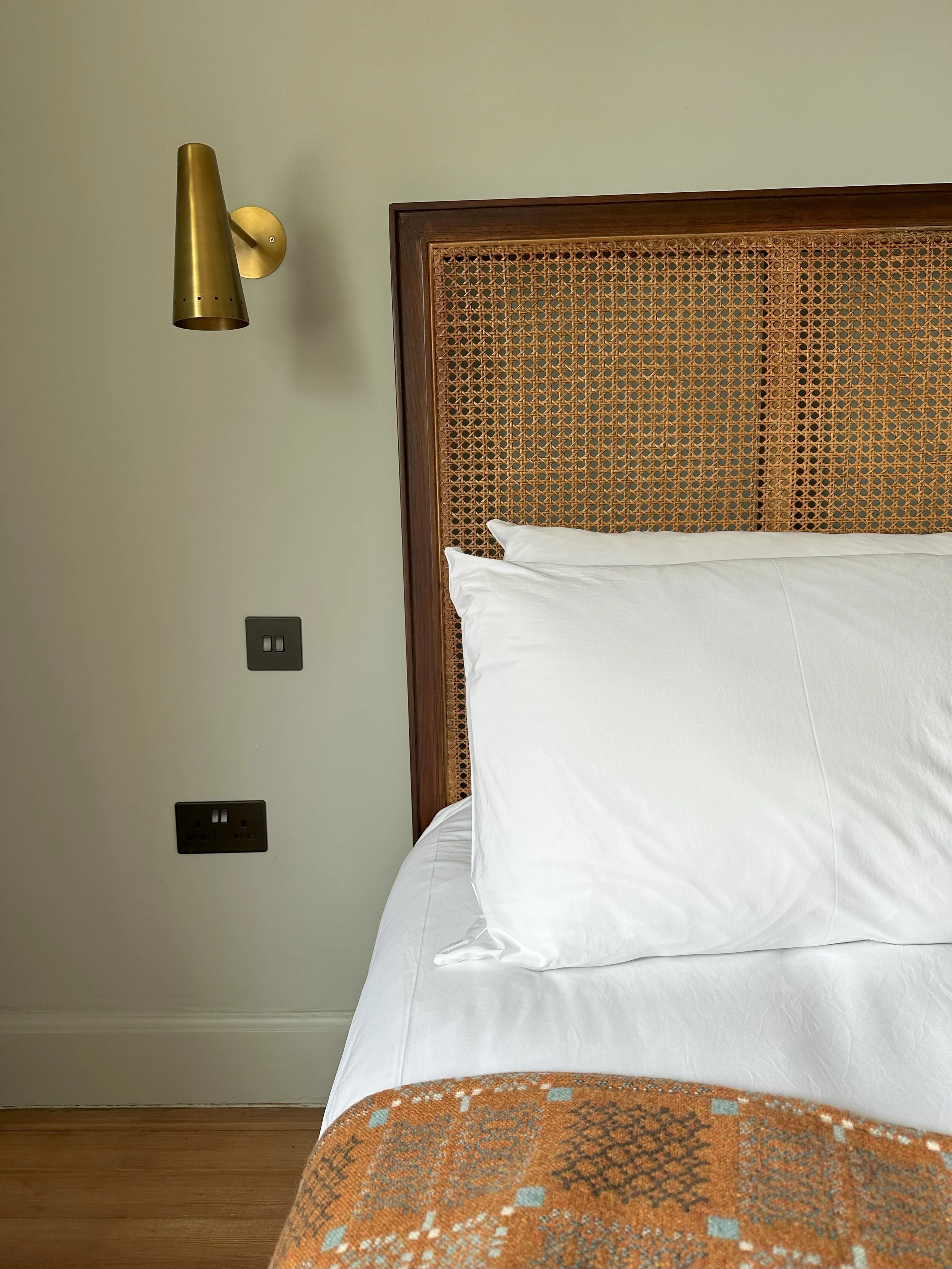 Close-up of a hotel bed with white pillows and a patterned orange and blue bedspread, wooden headboard, beige wall with mounted gold wall lamp and electrical outlets.