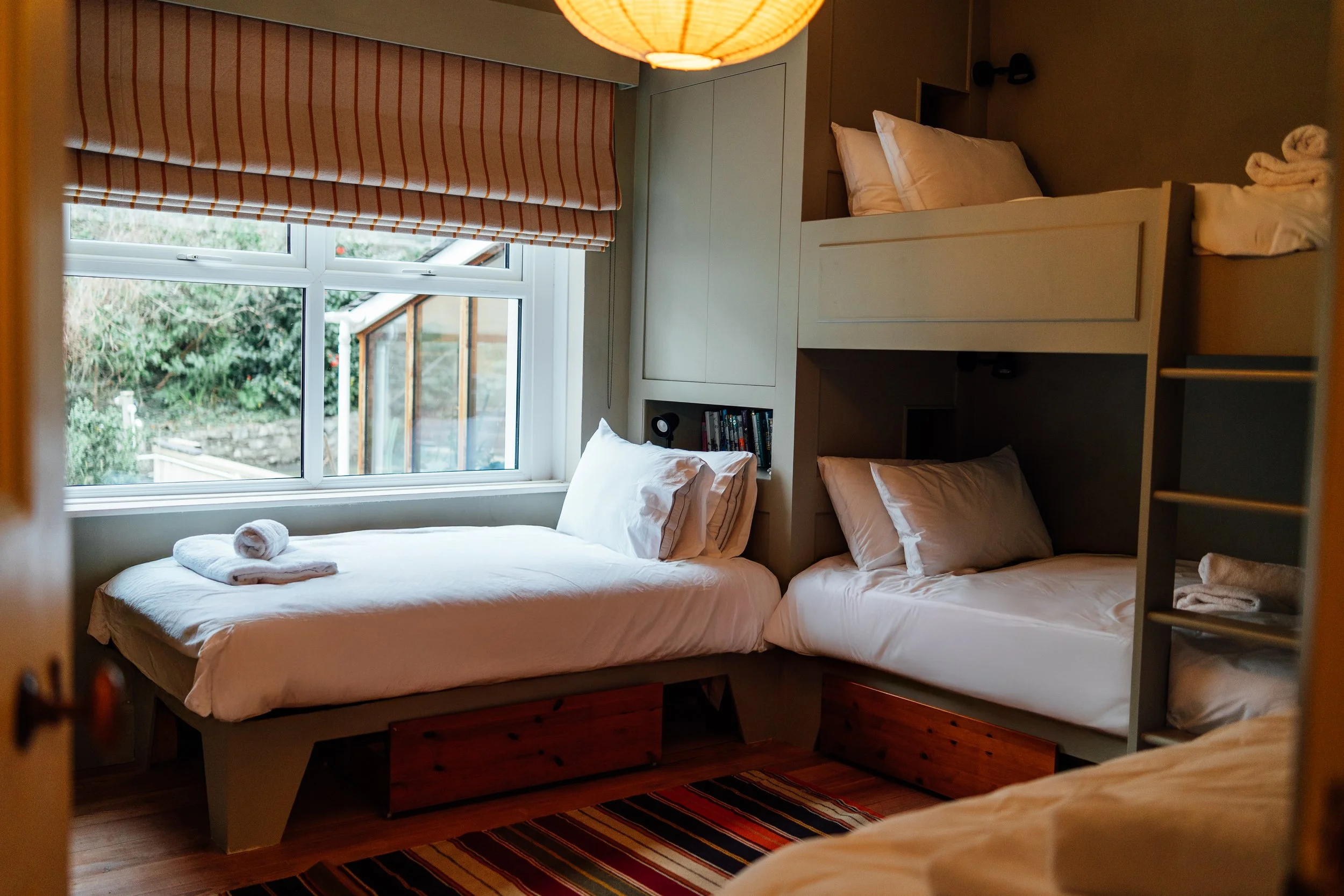 Room with two beds, one on the left near a window and a bunk bed on the right, with white linens and pillows, a window with curtains showing greenery outside, and a striped rug on a wooden floor.