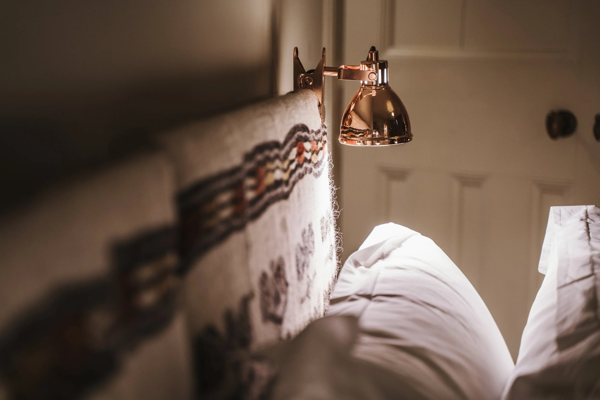 Close-up of a headboard with a wall-mounted copper reading light above a bed with white pillows.