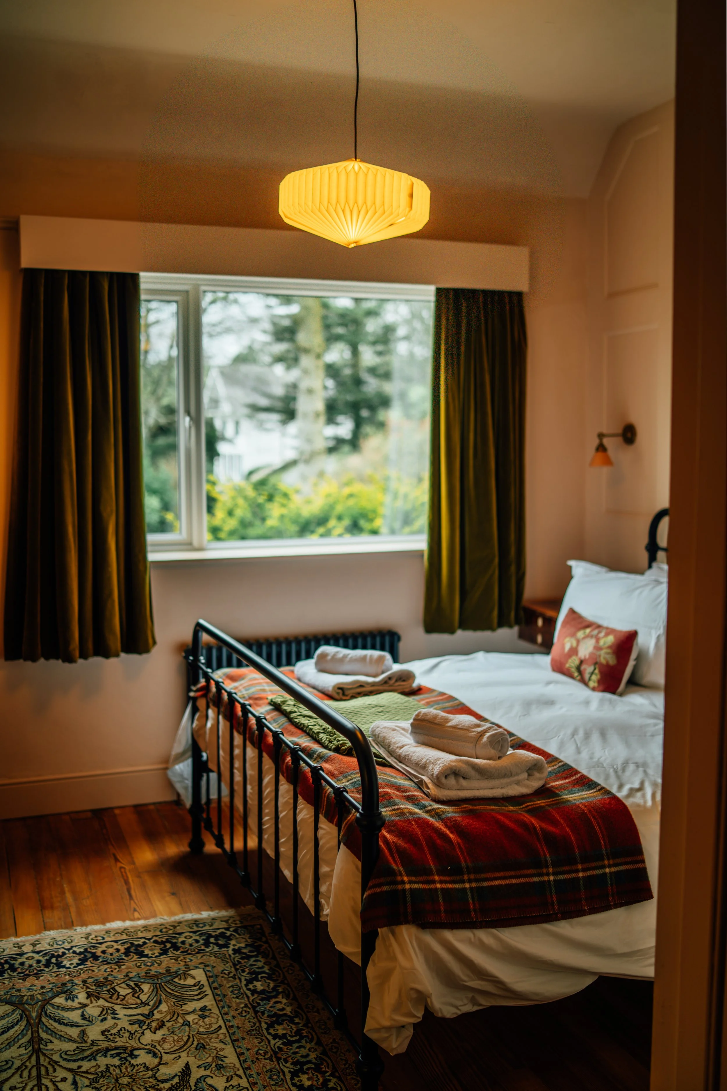 A cozy hotel room featuring a bed with white linens and a decorative pillow, dark green curtains, a large window with a view of trees, a plaid blanket at the foot of the bed, and neatly folded towels on top.