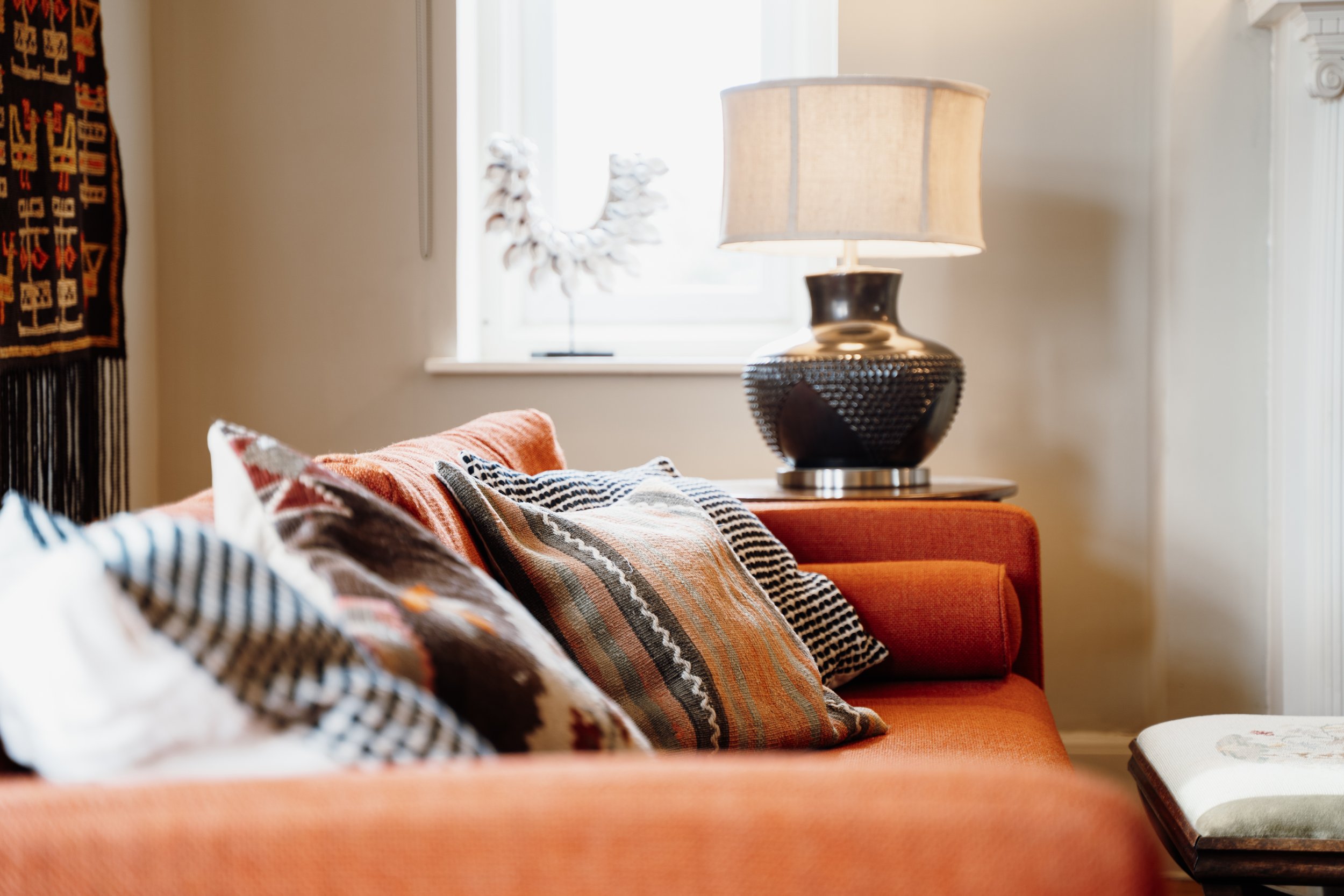 Living room with an orange sofa decorated with patterned cushions, a large black textured table lamp on a side table, a window with sunlight, and a decorative sculpture in the window. Part of a wall hanging is visible on the left.