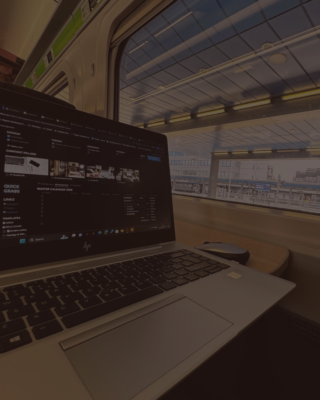 Laptop on a train table with a window view of train station platforms and overhead structures.
