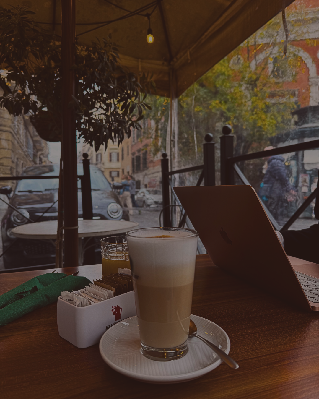 A cafe scene with a latte, sugar packets, and a laptop on a wooden table, with a street view through large glass windows showing cars and pedestrians.
