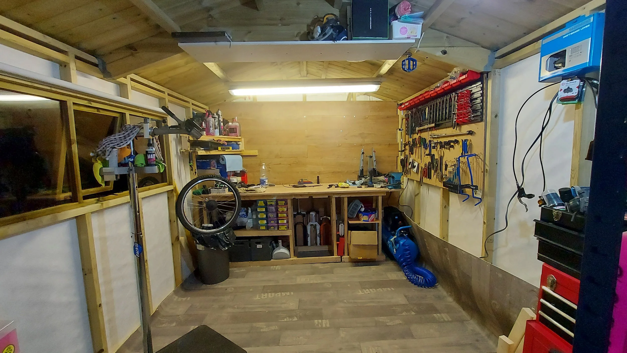 A well-organized woodworking or mechanical workshop with a wooden workbench, tools on the wall, shelves with supplies, a bicycle wheel hanging, and various equipment and containers around the space.