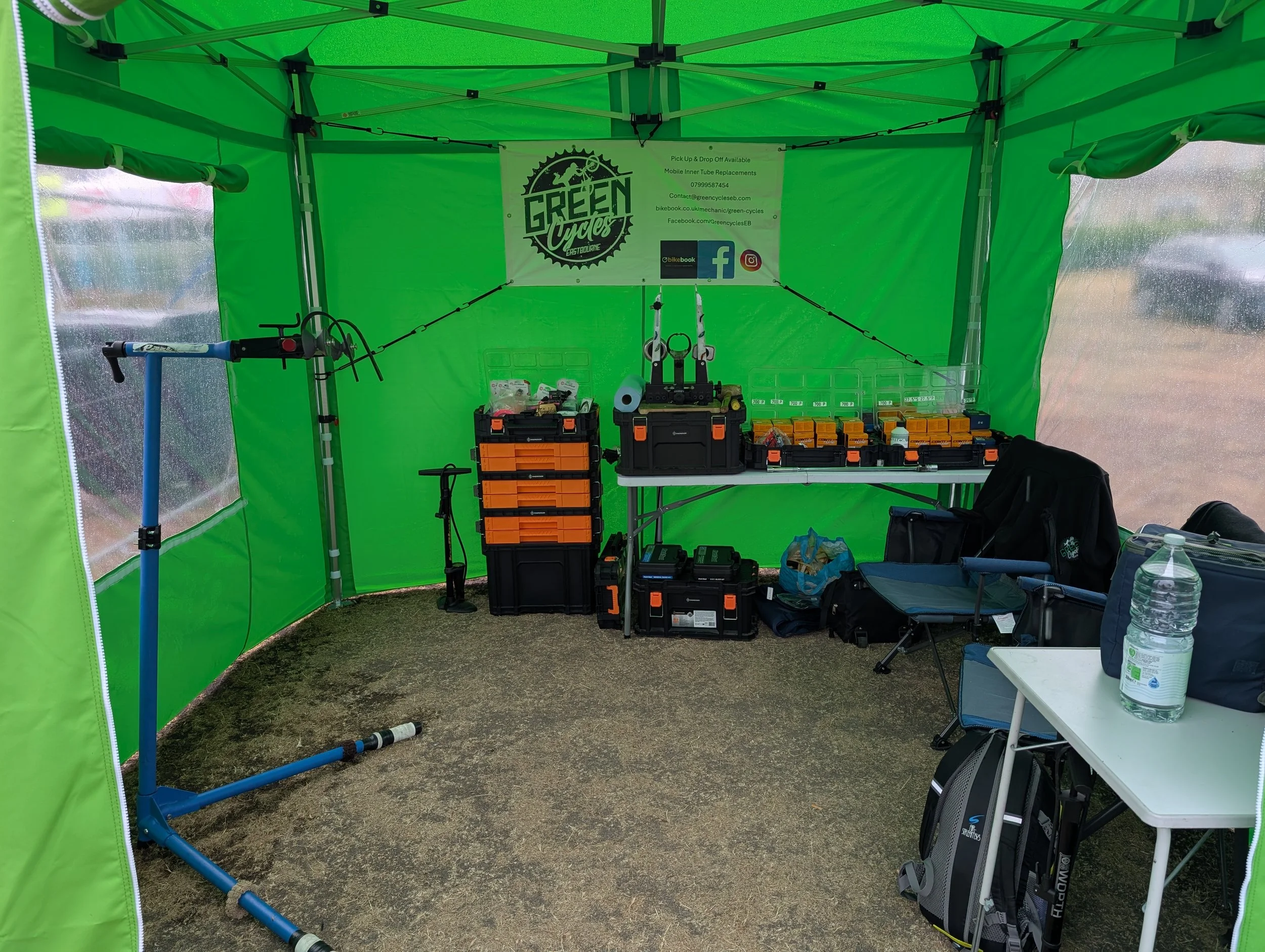 Green cycling booth with tools and equipment, a banner for Green Cyclery, and a large water bottle on table, set up outdoors on grassy ground.