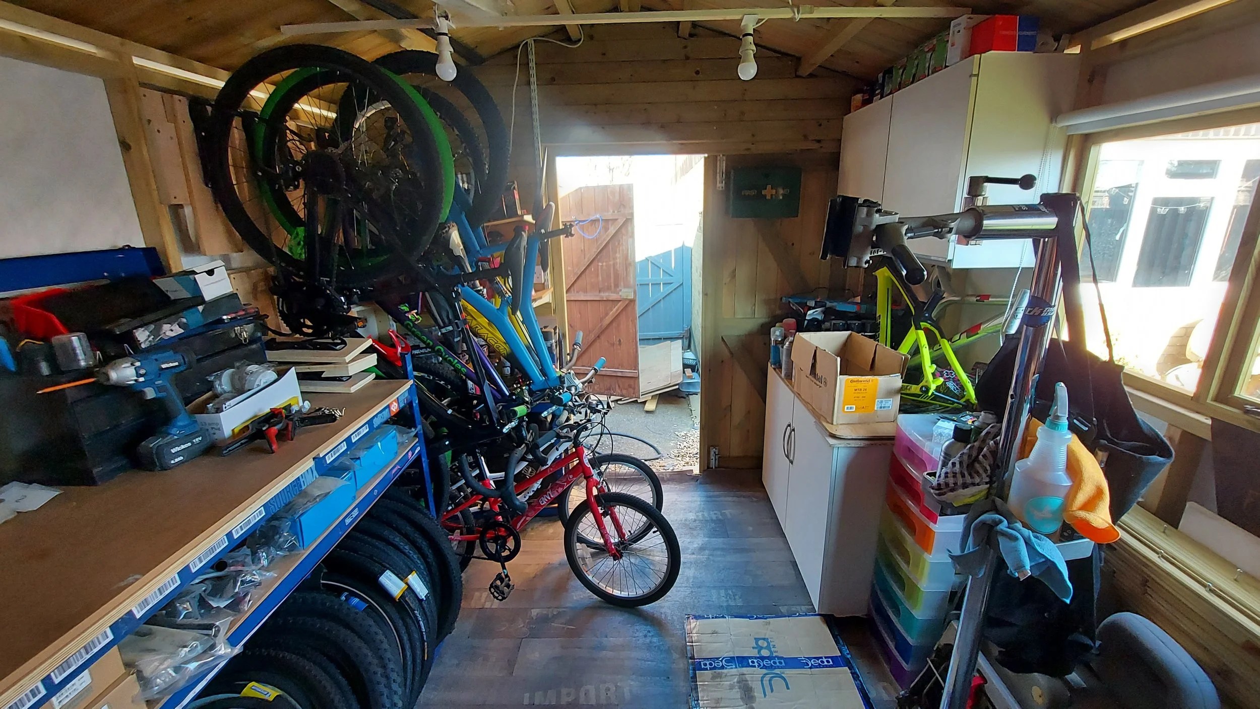 A cluttered garage with bicycles, tools, and storage containers, some bicycles stacked and leaning against the wall, a workbench with supplies, and a partially open door leading outside.