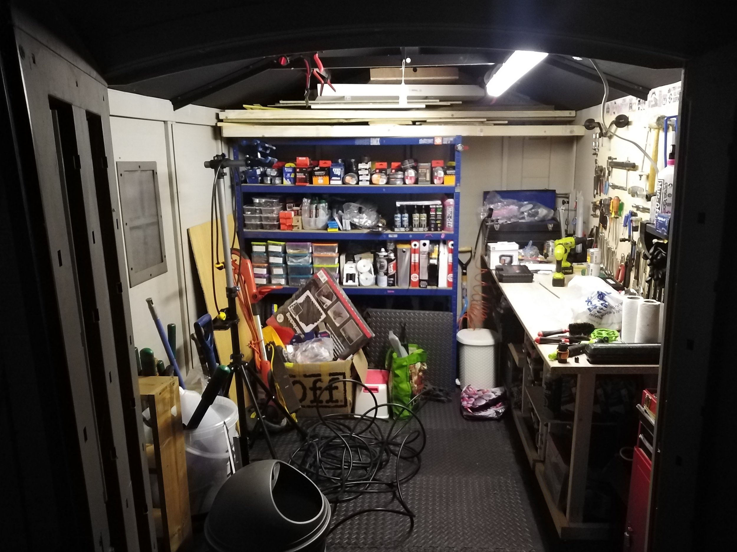 A cluttered workshop or garage with various tools, equipment, and supplies organized on shelves and workbenches, including power drills, small parts containers, and hand tools.