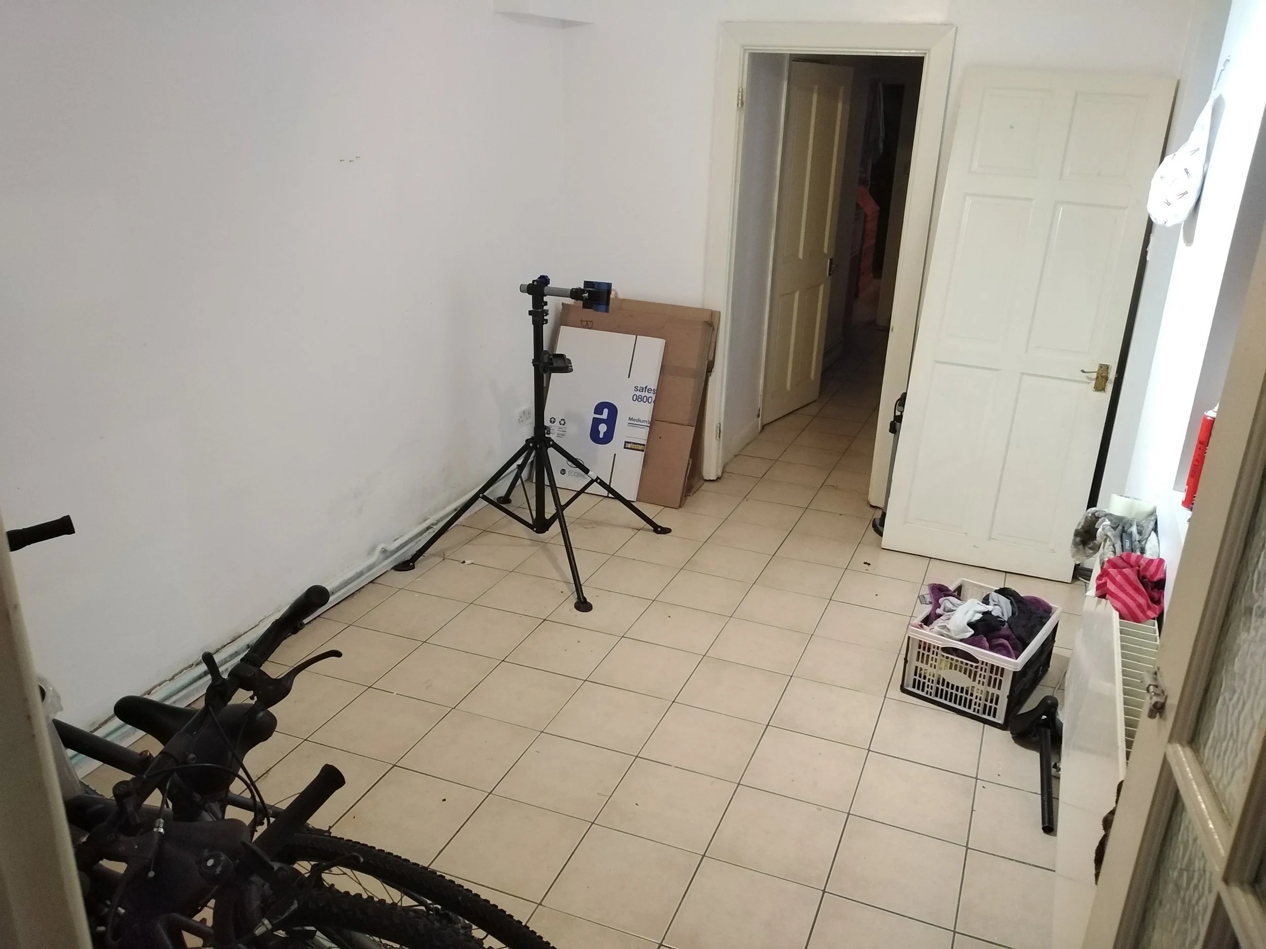 Empty room with open door, bicycle, tripod, boxes, laundry basket with clothes, and scattered items on the floor.