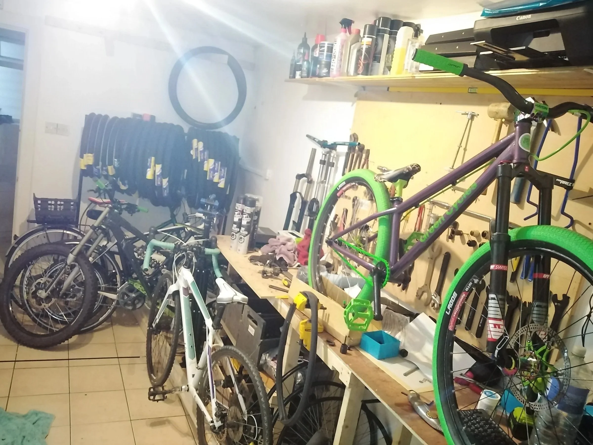 A cluttered workshop with multiple bicycles, tools, and supplies. There are two bicycles in the foreground, one above the other, one green and one white. In the background, a wall-mounted bike rack holds several tires. The workbench is covered with tools, parts, and cleaning supplies, with shelves above holding bottles and spray cans.