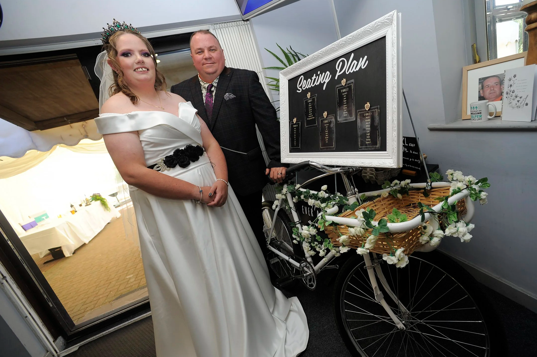 Bride in white wedding dress and tiara standing with groom in a dark suit next to a seating plan display on a decorated bicycle.