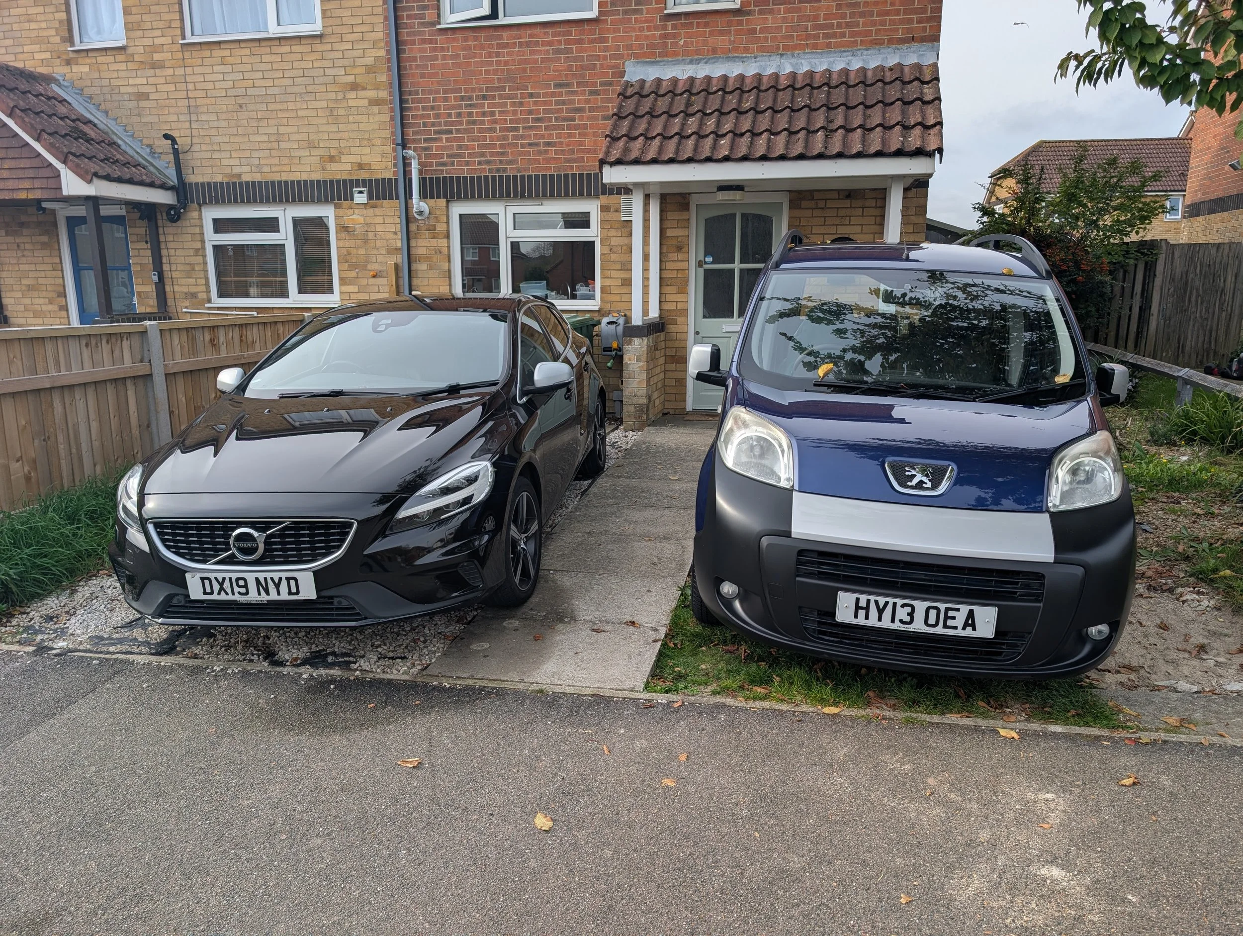 A driveway with two parked cars in front of a house. The car on the left is a black Volvo with a license plate DX19 NYD, and the car on the right is a blue Peugeot with a license plate HY13 OEA. The house has a small porch with a door and window, surrounded by a wooden fence on the left and a garden on the right.