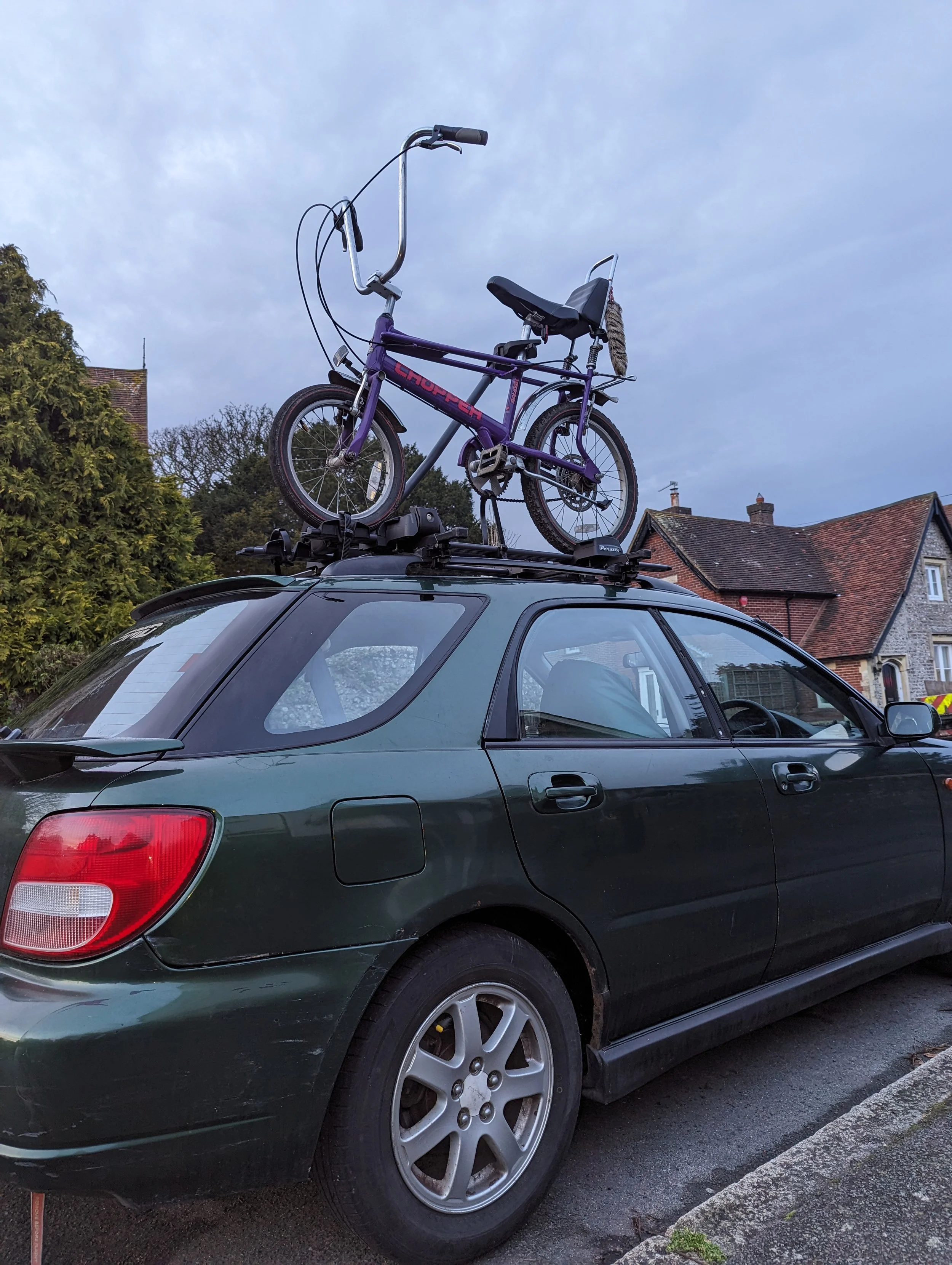 Green car with a bicycle mounted on the roof, parked on a residential street during evening, with houses and trees in the background.