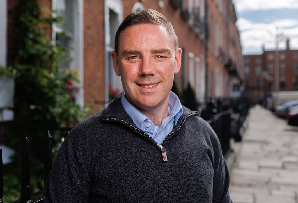 Patrick O'Rourke, a certified career coach, standing on a sidewalk in an urban neighborhood with brick buildings and parked cars in the background.