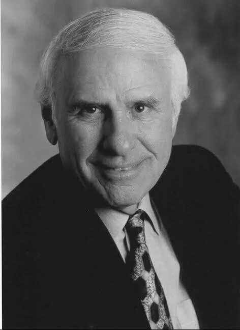 Black and white portrait of an older man with white hair, wearing a dark suit jacket, a collared shirt, and a patterned tie, smiling slightly.