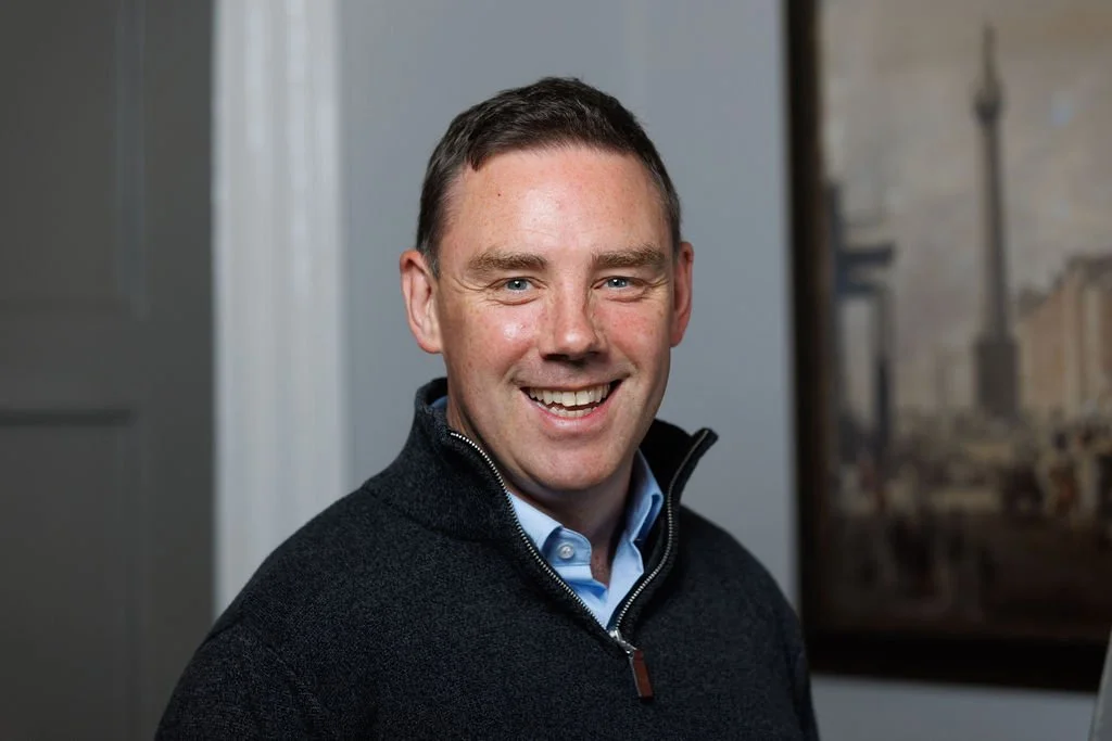 A smiling man with short dark hair wearing a dark zip-up sweater over a light blue collared shirt, standing indoors near a wall with a framed picture in the background.