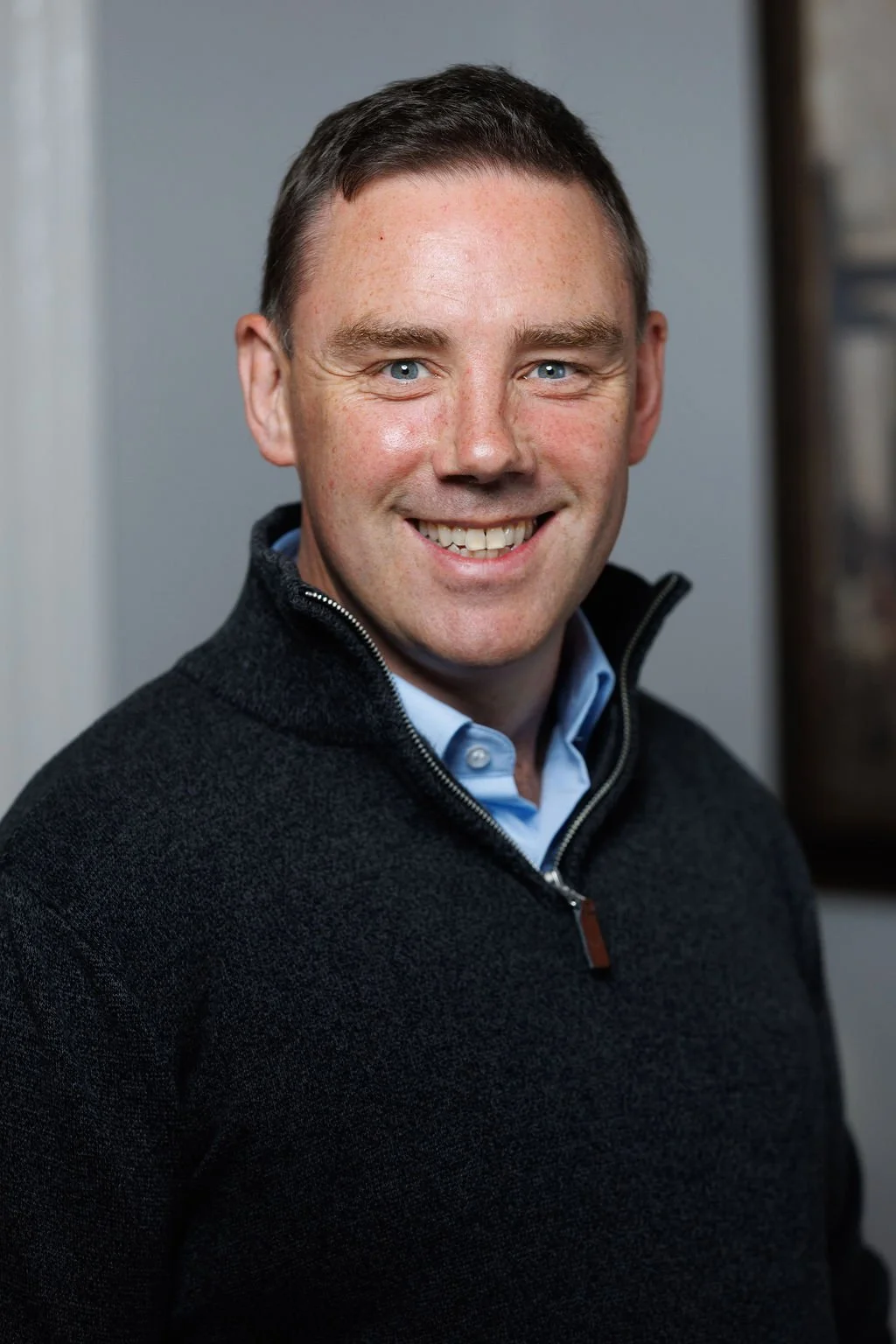 A smiling man with light skin, short brown hair, and blue eyes, wearing a dark zip-up sweater over a light blue collared shirt, standing indoors with a plain gray background.