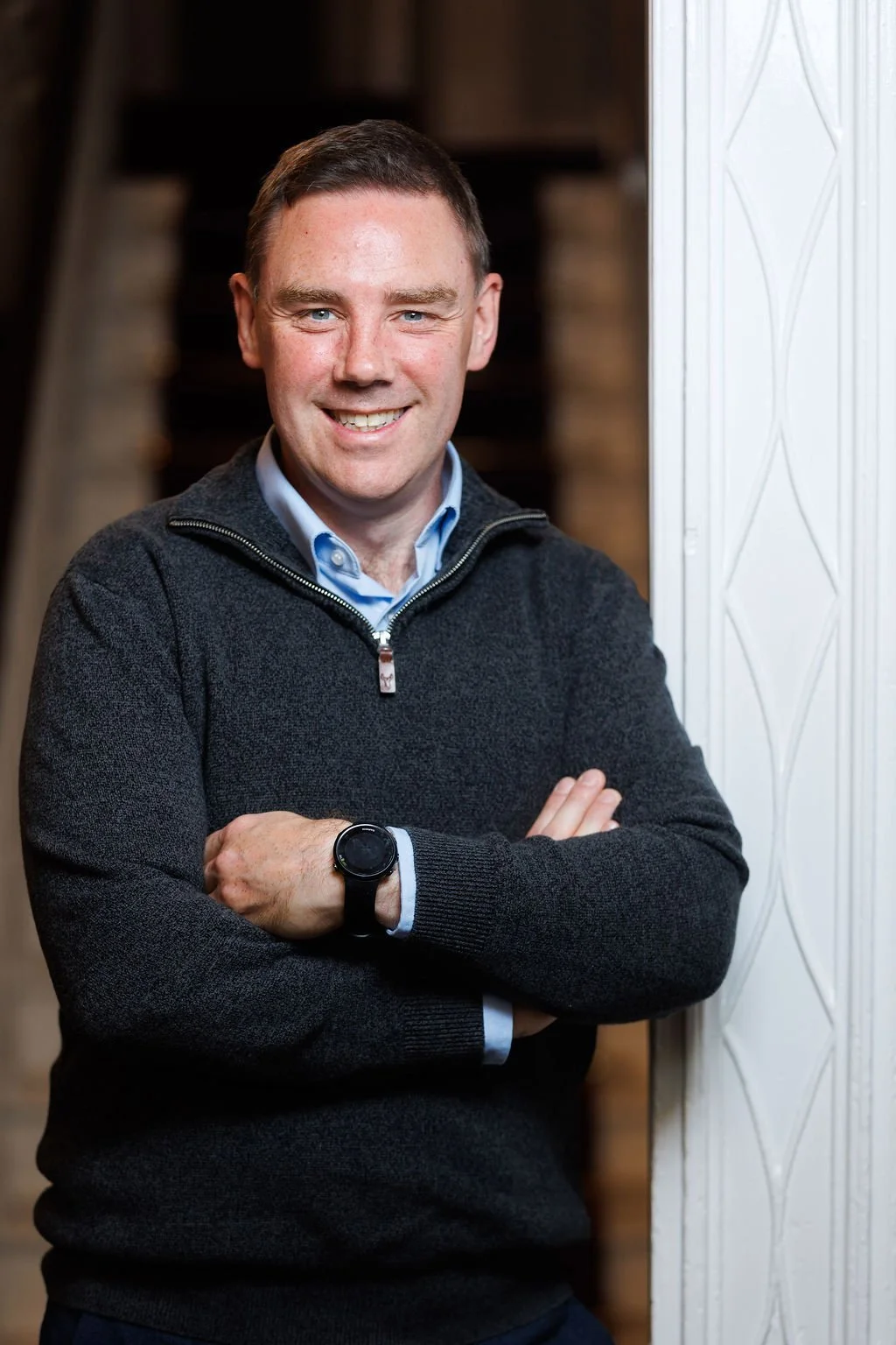 Patrick O'Rourke, a certified career coach standing with arms crossed, wearing a black zip-up sweater, blue shirt, and a smartwatch, standing next to a white textured wall with stairs in the background.