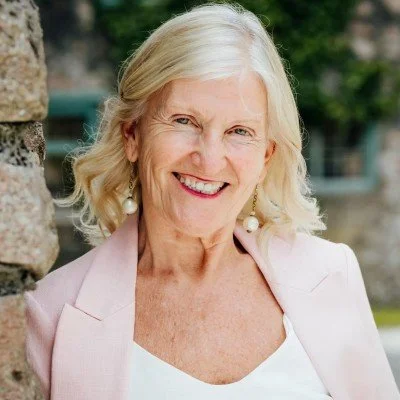 A smiling elderly woman with blonde hair, wearing a light pink blazer and white top, standing outdoors near a stone wall.