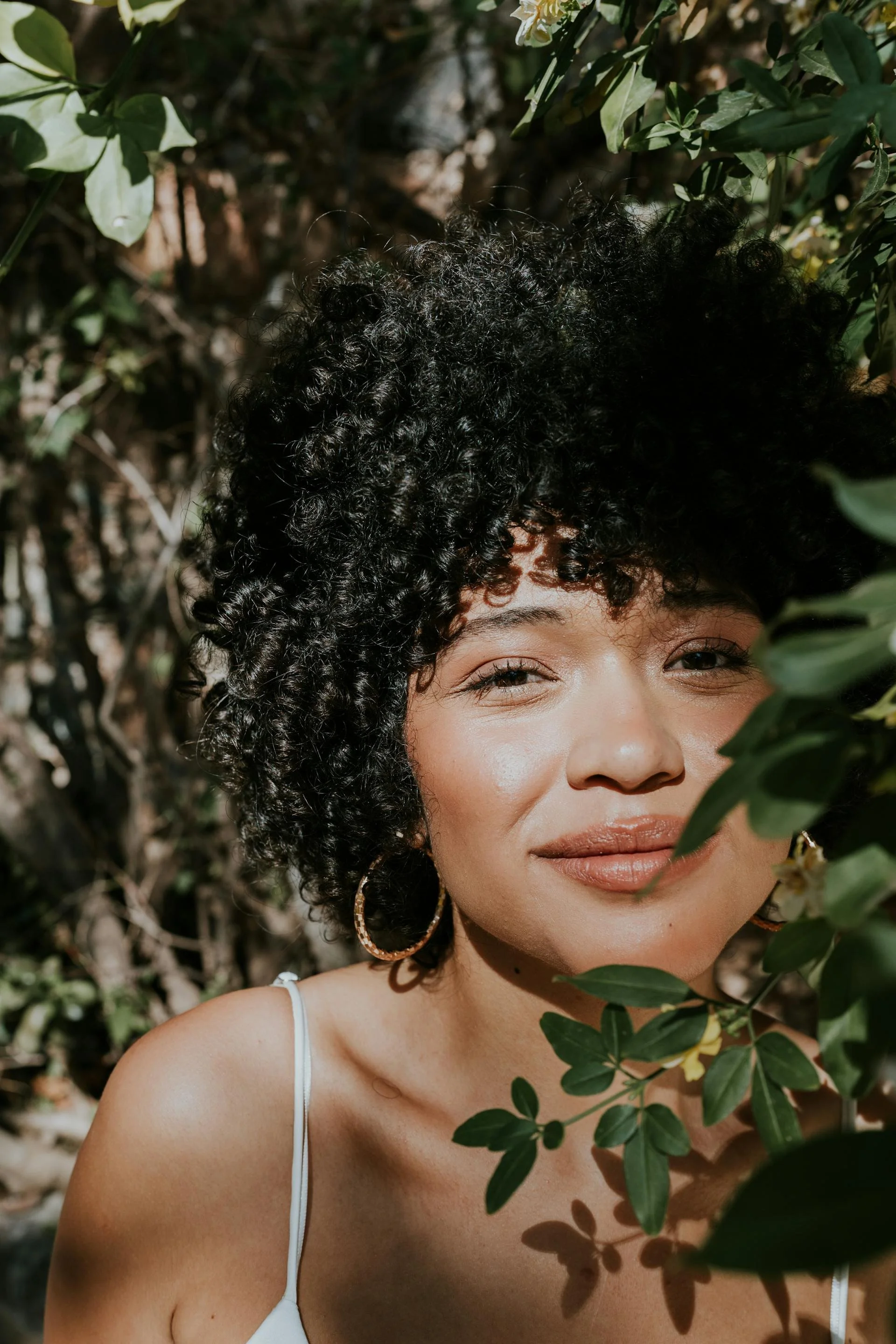a softly smiling Black woman looking out from behind dark green leaves.