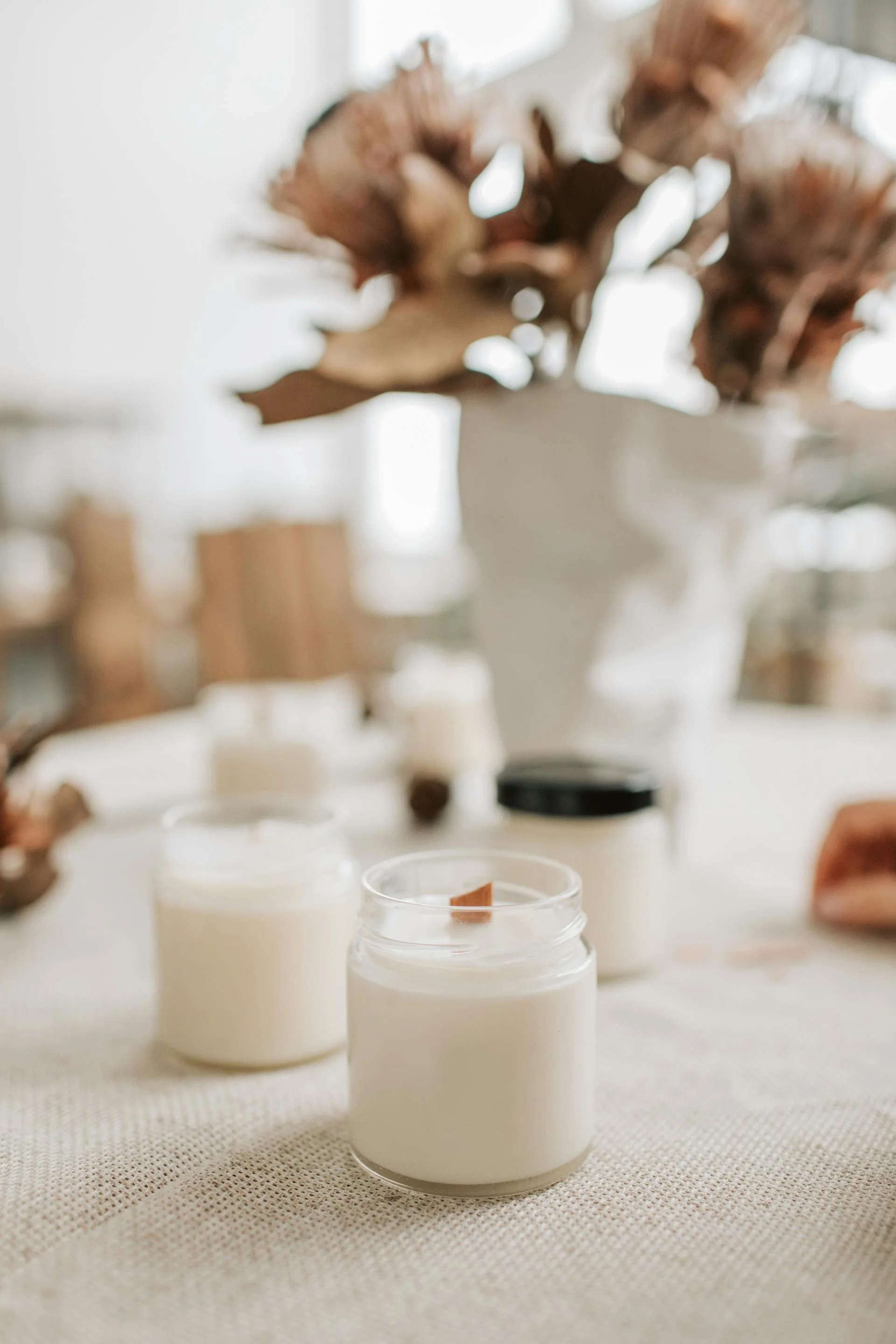 Close-up of white candles on a beige textured tablecloth, with a blurred background of a vase with dried flowers and other objects.
