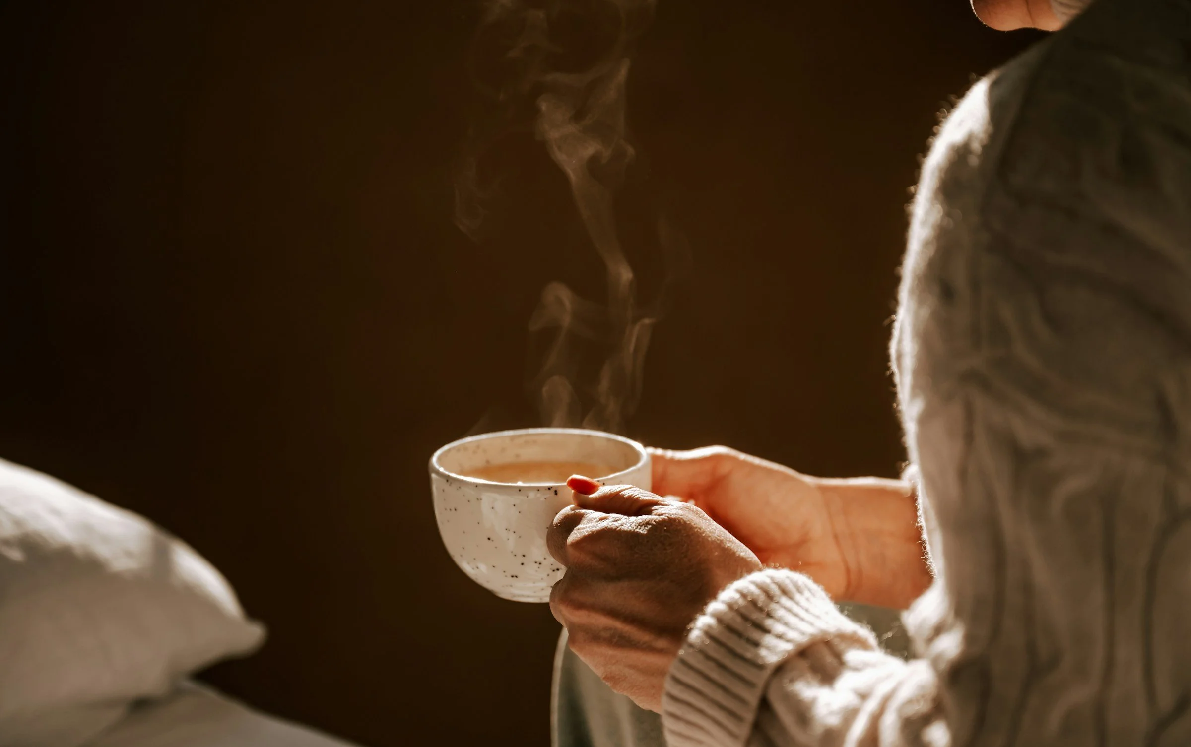 Person holding a steaming cup of hot beverage in a cozy setting.