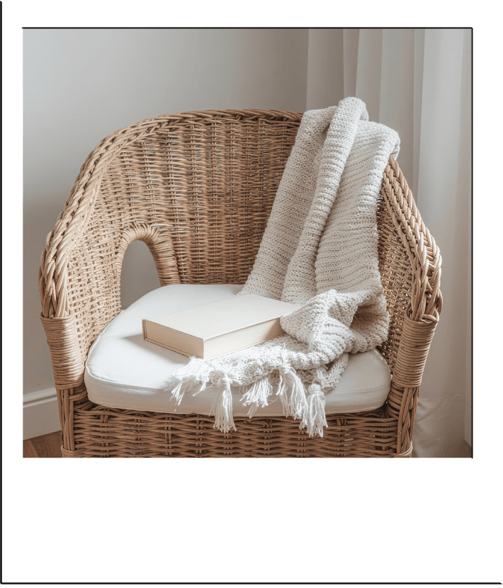 A wicker chair with a white cushion, a beige book, and a cream-colored knitted throw blanket draped over it.