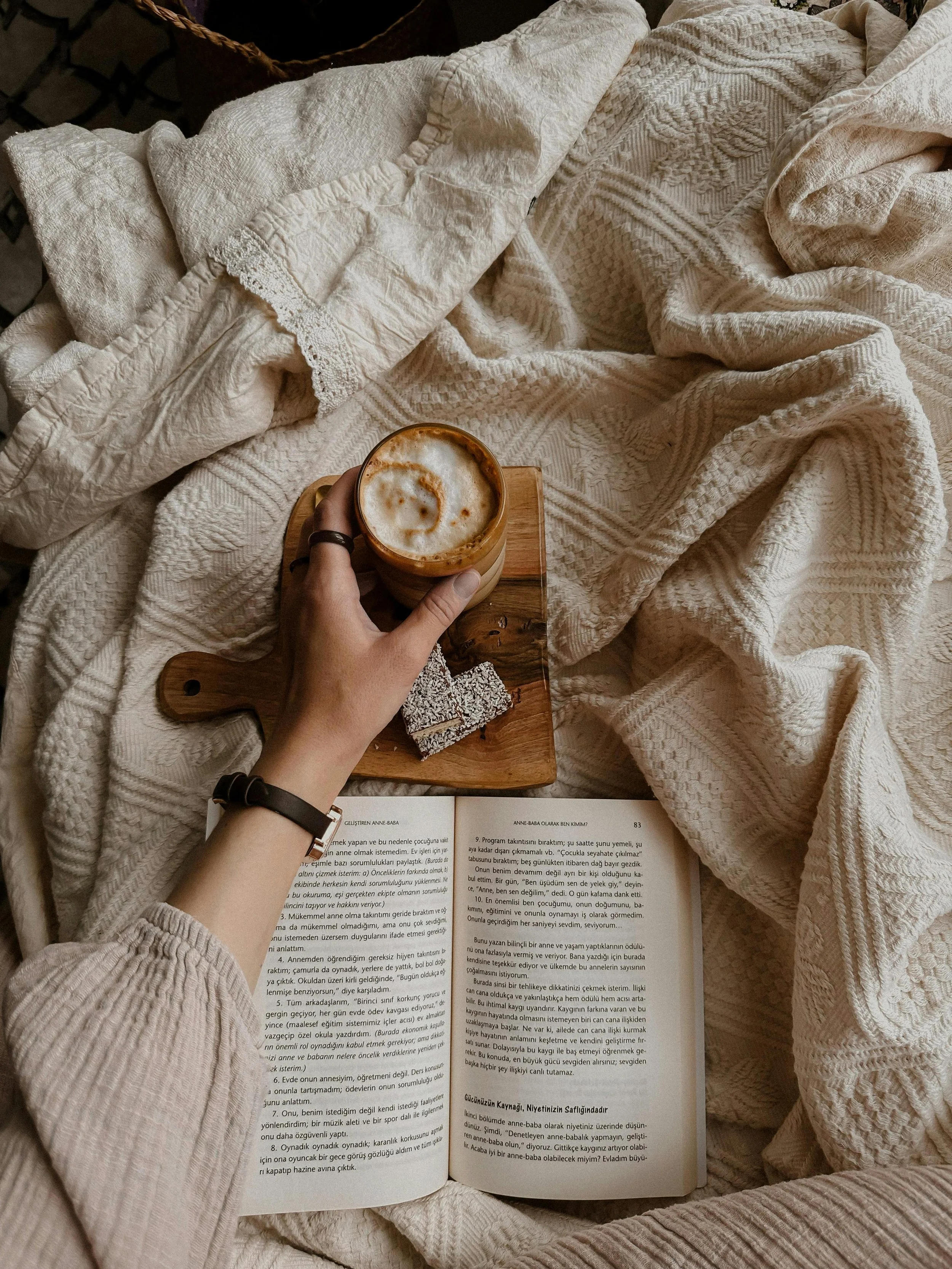 overhead view of person holding a foamy mug with open book and a blanket.
