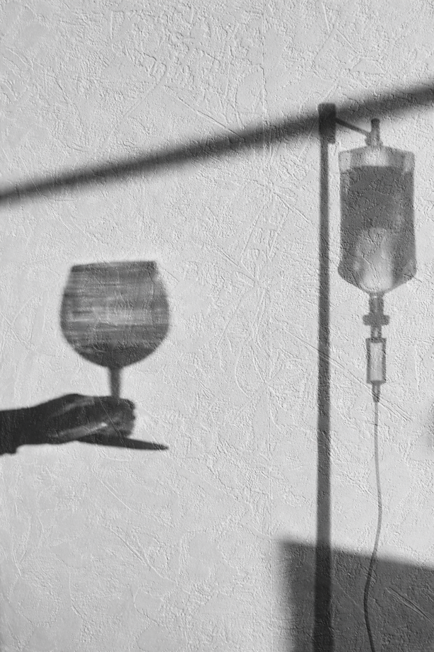 A shadow of a hand holding a wine glass cast on a textured wall, with a shadow of an IV drip on the wall.