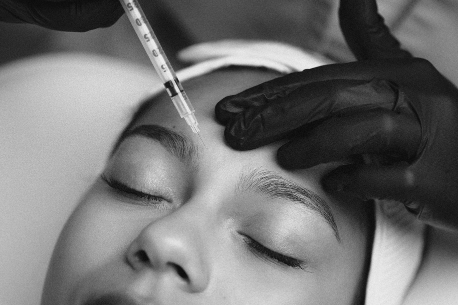 A person receiving a cosmetic treatment on their eyebrows using a syringe, with a practitioner wearing black gloves.