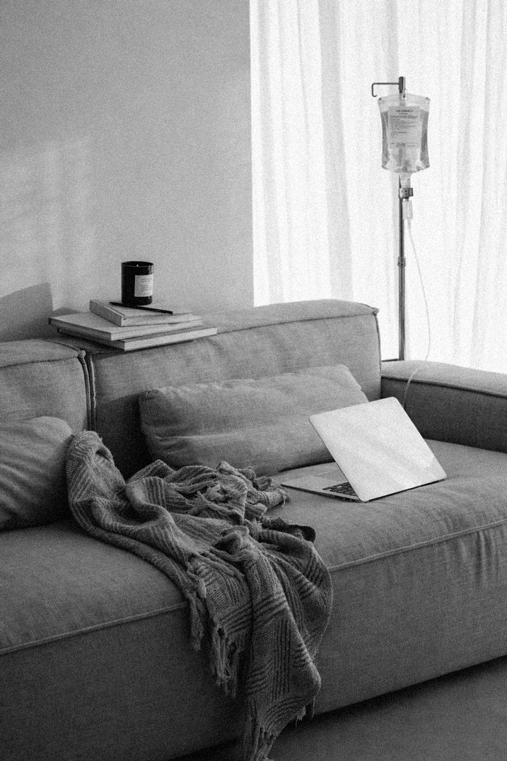 A living room with a beige sofa, a blanket, a laptop, and a pillow. There is a stack of books with a jar on top on the backrest. An IV stand with a bag hangs near a curtain in the background.