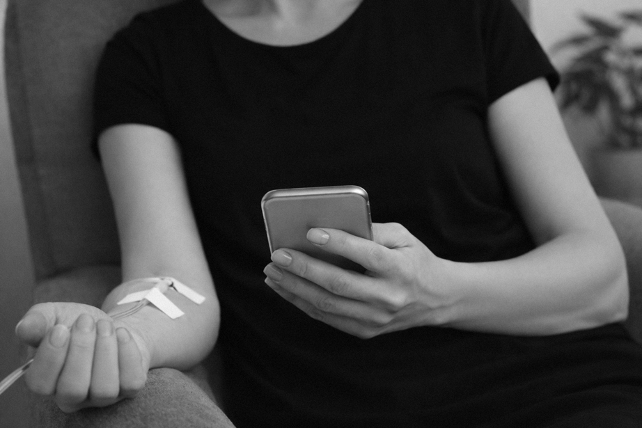 A woman in a black shirt is sitting on a couch, holding a smartphone in her right hand and her left arm extended with a needle inserted into her arm, connected to an IV tube.