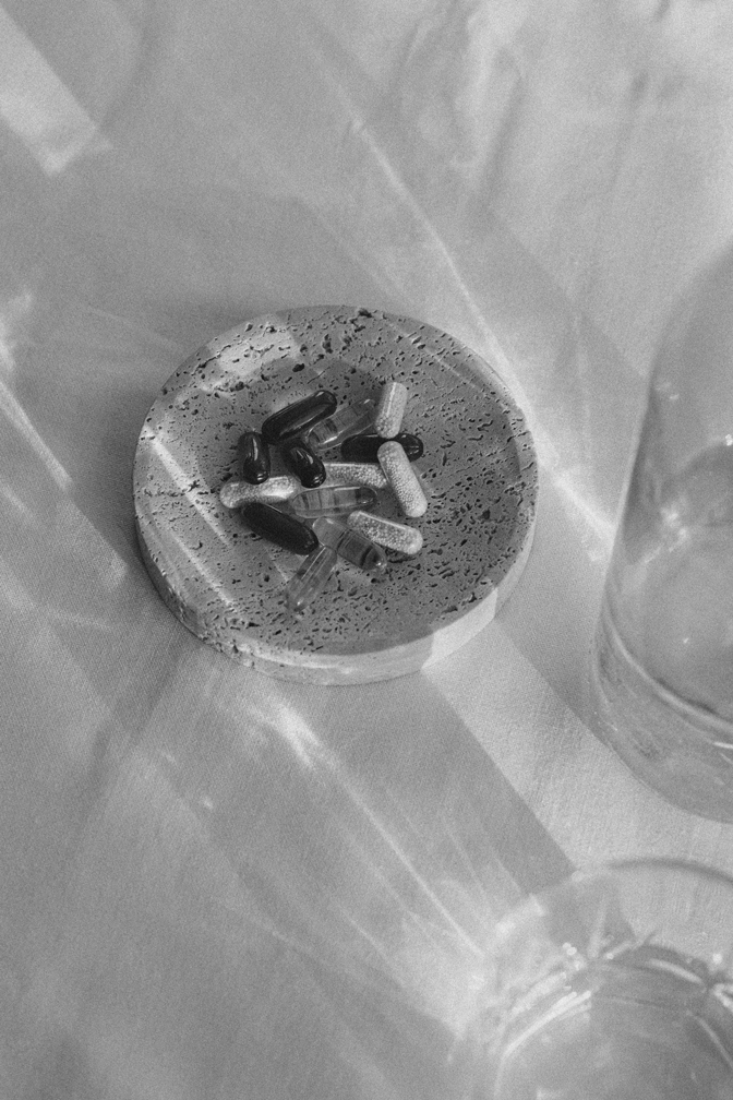 A round soap dish with various capsule pills on it, next to a glass of water on a table.