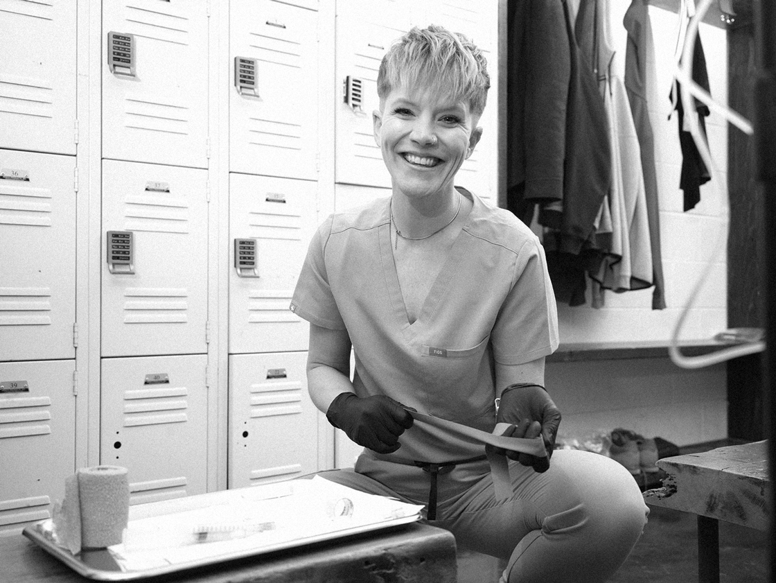 A smiling woman in medical scrubs and gloves sitting in a locker room.