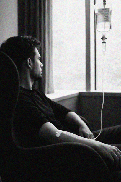 A man sitting in a chair with an IV drip attached to his arm, looking out a window.