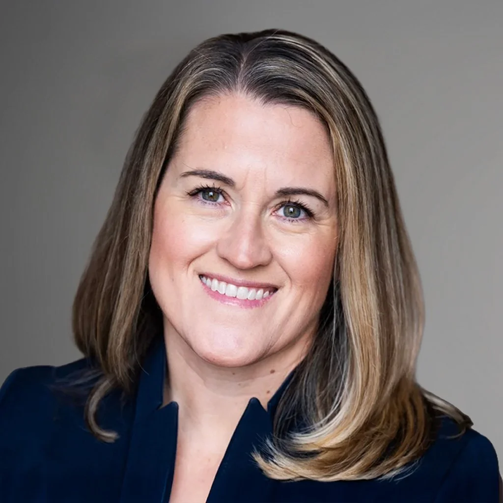 A woman with shoulder-length light brown hair and blue eyes smiling at the camera, wearing a dark blazer against a neutral background.