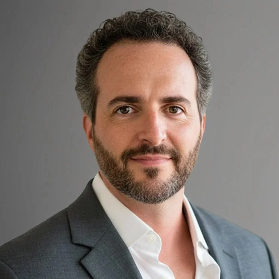 A professional man with curly dark hair, beard, and mustache, wearing a dark suit jacket and white shirt, against a plain gray background.