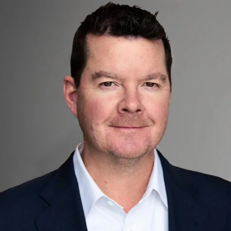 Professional headshot of a man with short dark hair, wearing a dark suit jacket and white dress shirt, smiling slightly against a gray background.