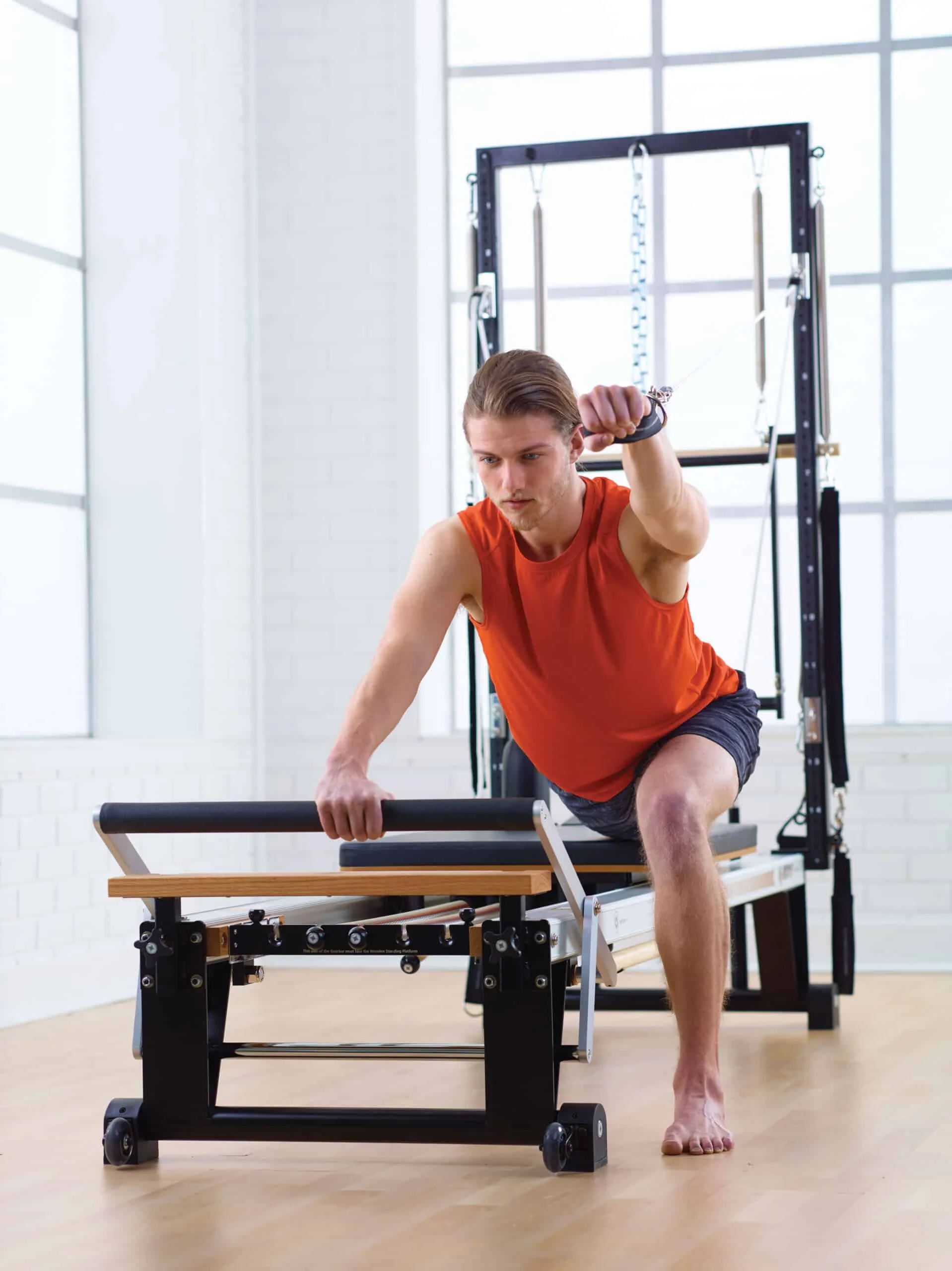 A man with long hair in an orange sleeveless shirt and shorts practicing Pilates on a reformer machine in a bright, spacious studio with large windows.