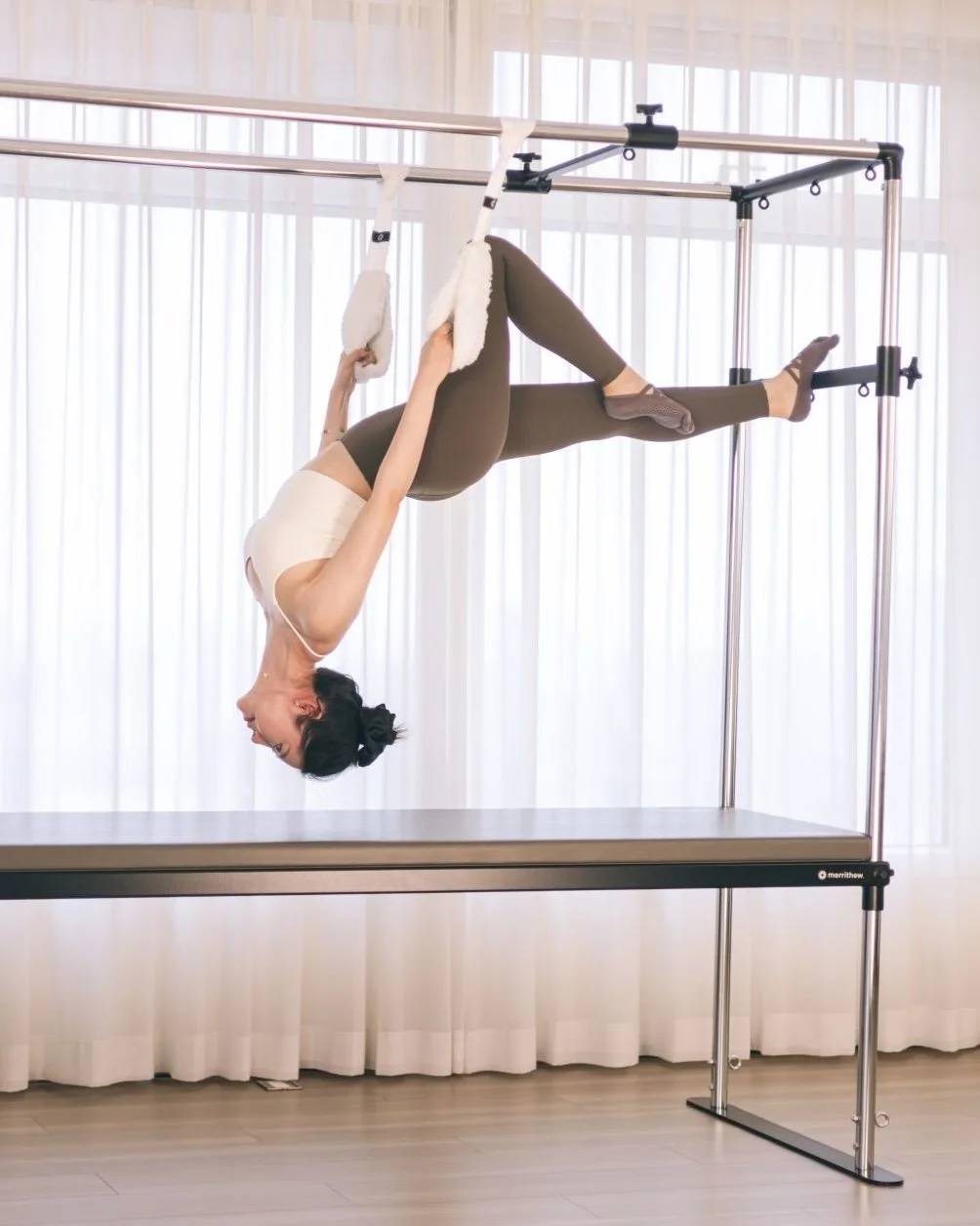 A woman practicing aerial silk yoga upside down on a metal frame, with her legs extended outward and holding onto the fabric with her hands, in a room with white curtains.
