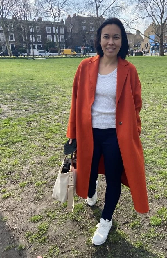 A woman standing outdoors in a park, wearing an orange coat, white sweater, blue pants, and white sneakers, holding a beige handbag.