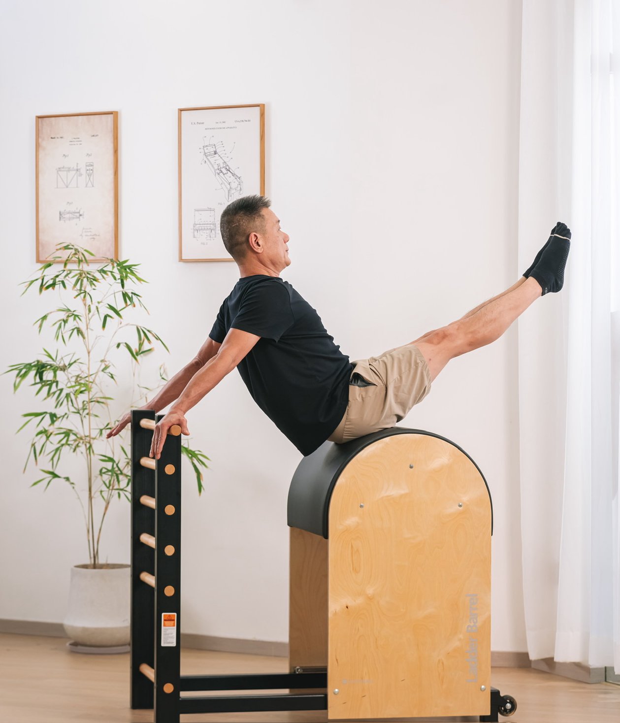 A man exercising on a Pilates chair, with his legs extended upward and his hands gripping handles, in a room with framed diagrams on the wall and a potted plant.