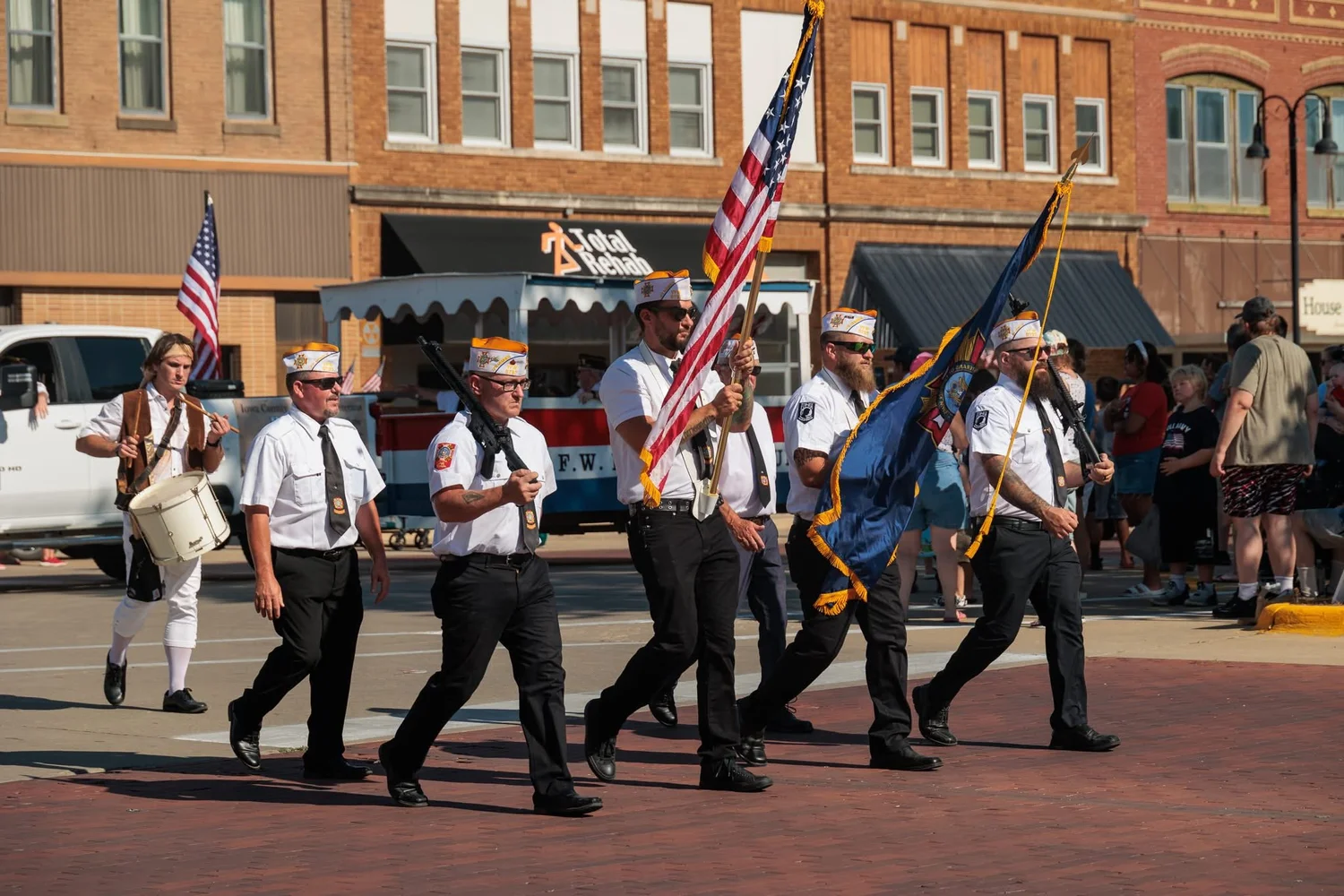 Group of veterans walking in a parade, holding American flags and a blue flag, dressed in white shirts, black pants, and hats, with a woman playing a drum in the background.