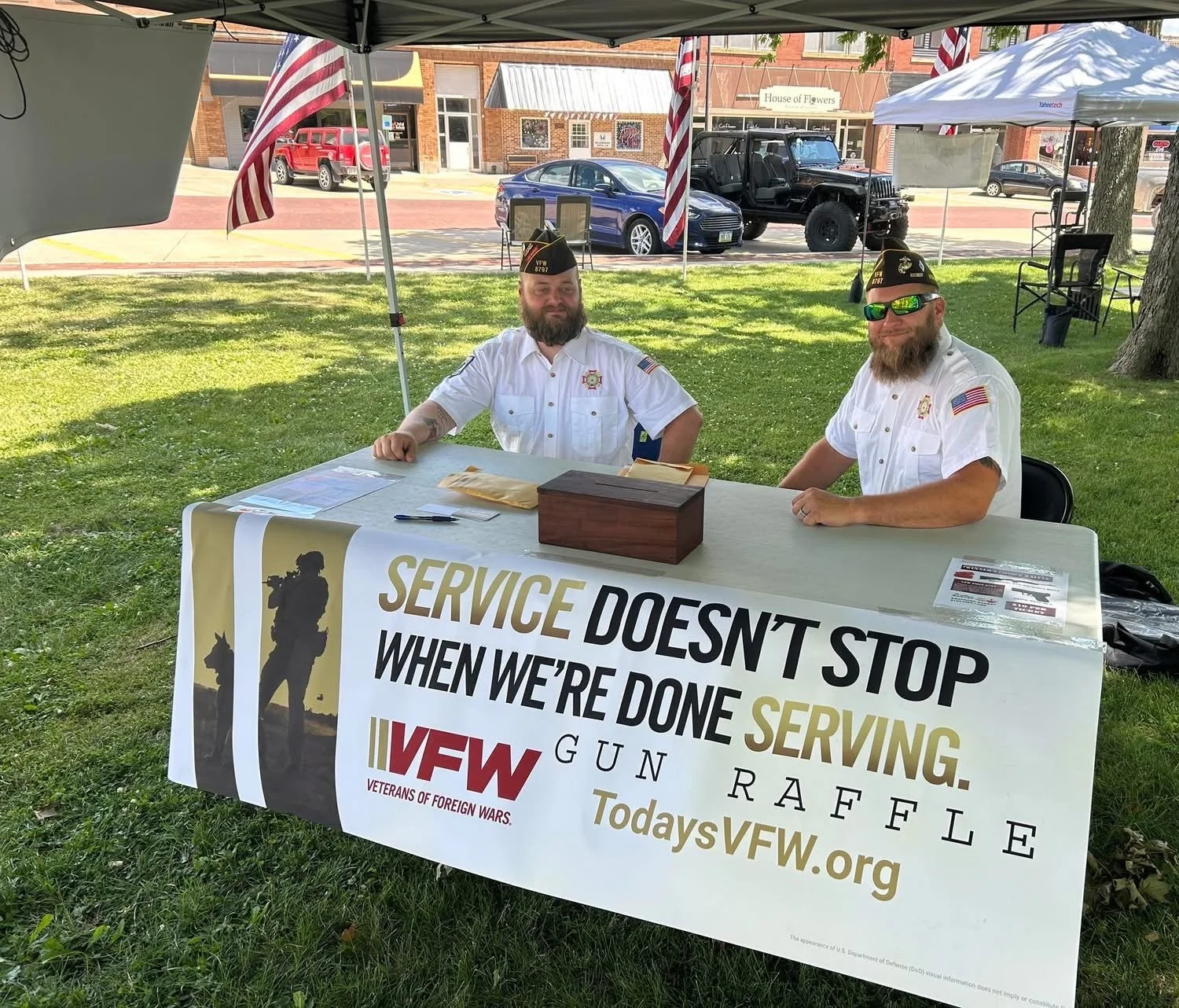 Two men in white veteran shirts and hats sitting at a table under a canopy with American flags, a sign reading 'Service Doesn't Stop When We're Done,' and a website URL, outdoors in a park-like setting with cars and stores in the background.