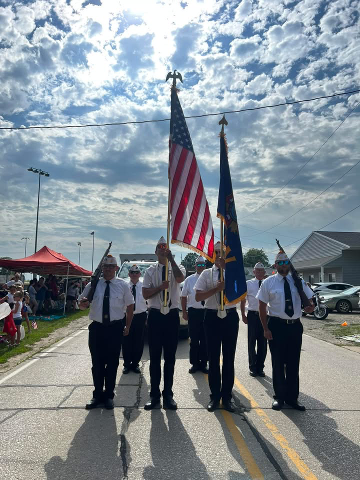 A group of people dressed in white shirts, black ties, and black pants marching in a parade holding American and state flags. Spectators sit under tents on the side of the street, and a house and parked cars are visible in the background under a partly cloudy sky.