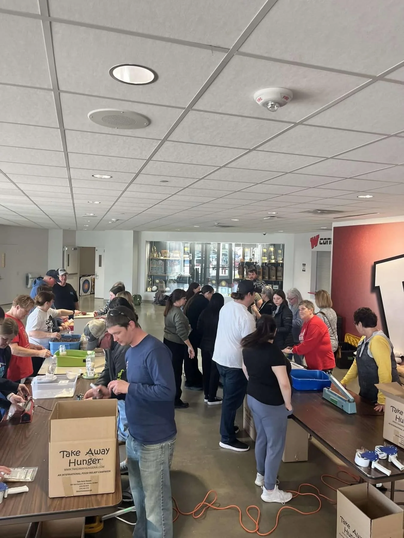 People lining up at a donation or charity event inside a building, some packing food or supplies at tables, with boxes labeled 'Take Away Hunger'.