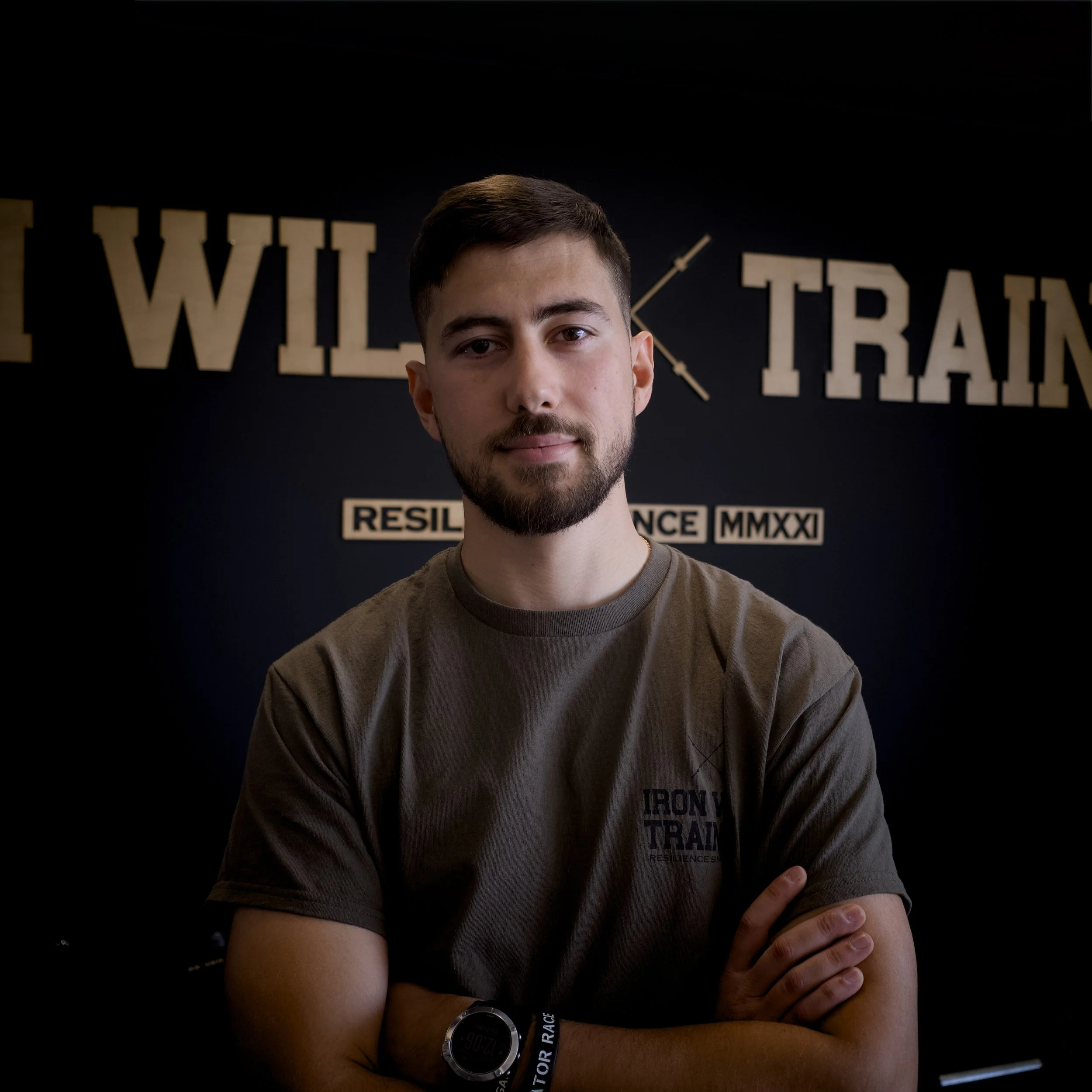 A young man with short dark hair, beard, and mustache, standing in front of a dark background with words and images related to strength training, including the text 'WILL' and 'TRAIN,' along with a clock illustration and phrases like 'RESILIENCE' and 'MMXXI.' He is wearing a brown T-shirt with text and an athletic wristwatch, and has a confident expression with arms crossed.